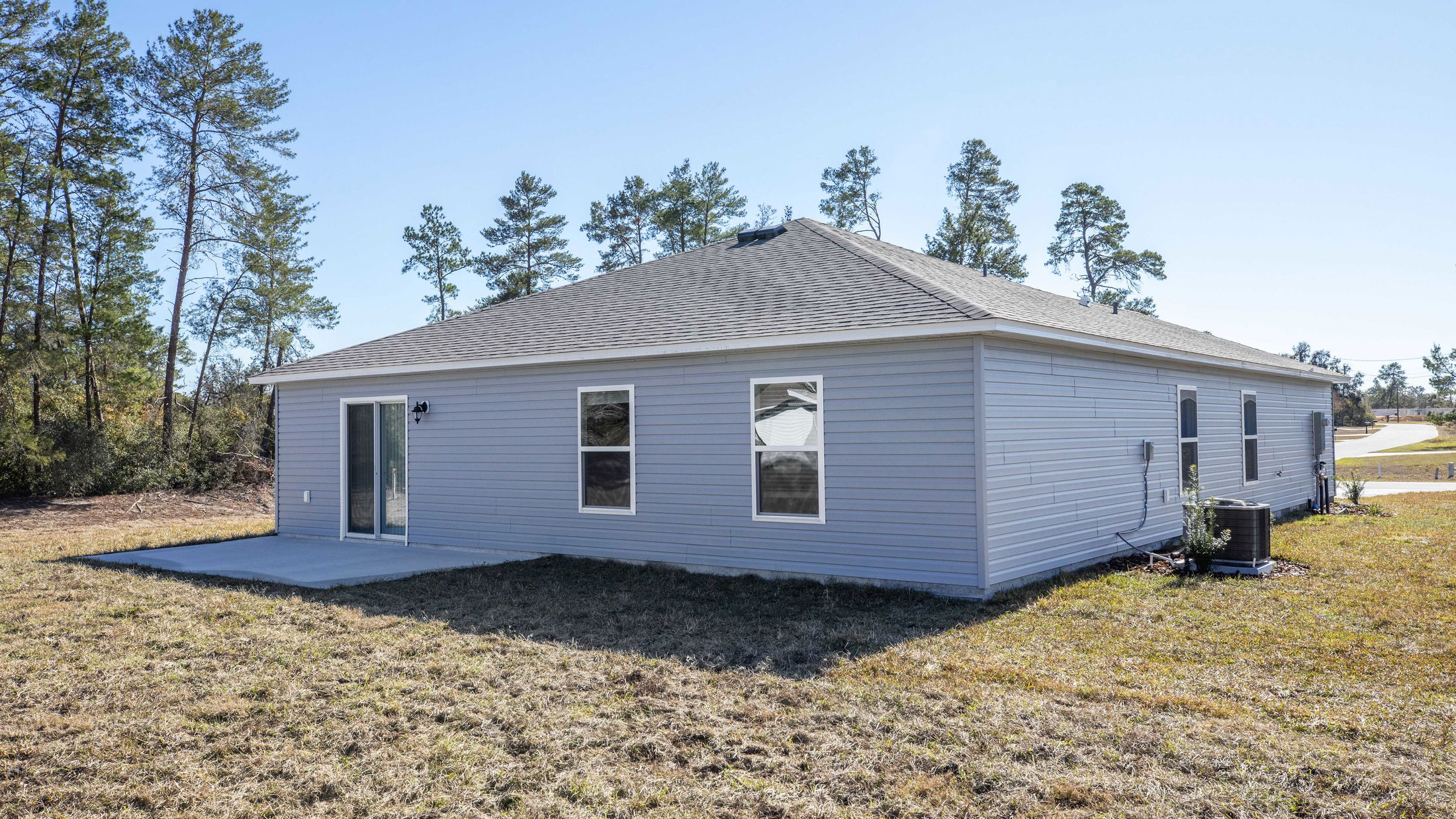 Rear view of a modern, single-story gray house with a sloped roof, surrounded by a grassy yard and trees under a clear blue sky.