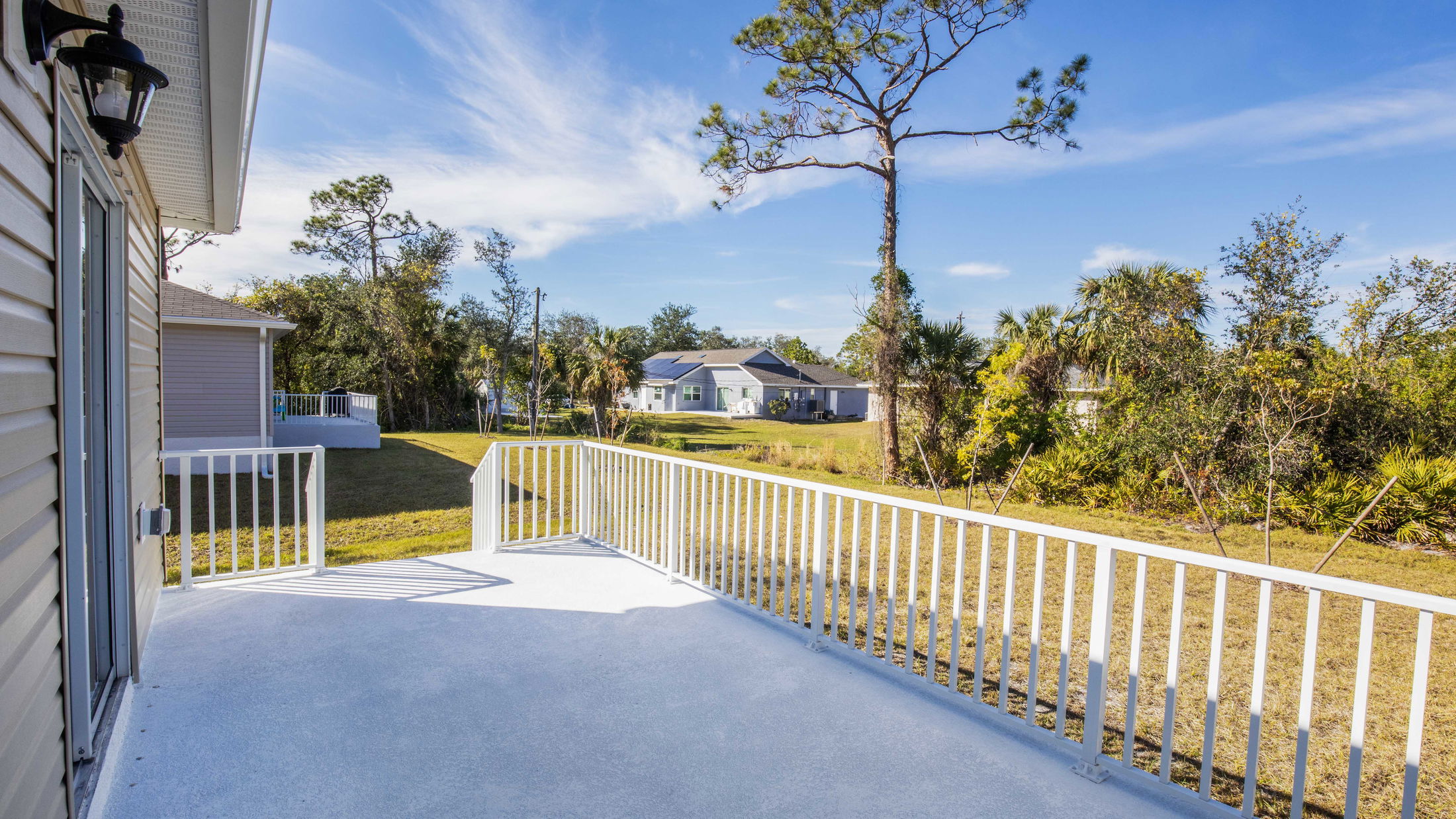 Spacious backyard view with trees and a white railing veranda leading to neighboring houses under a clear blue sky.