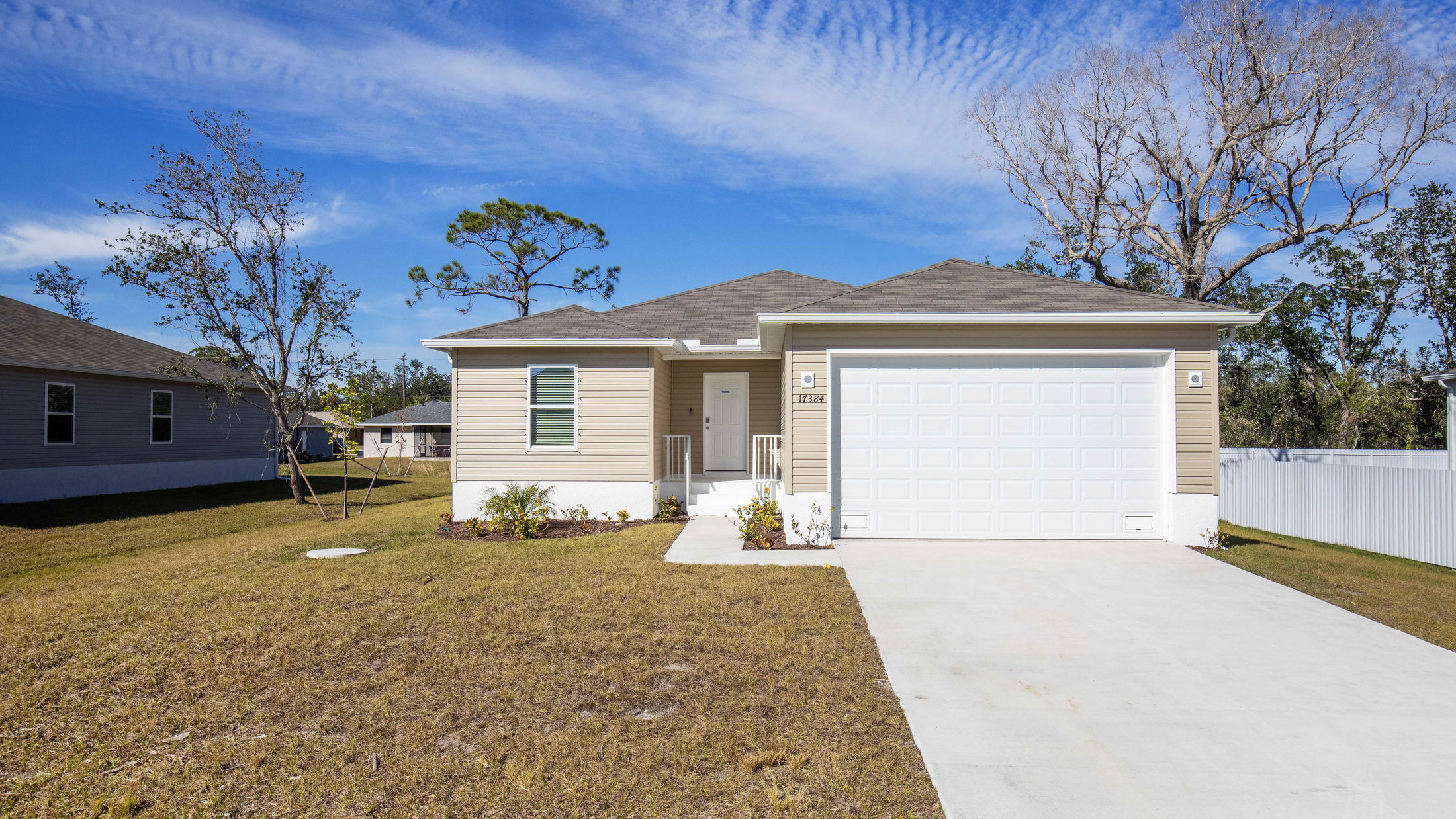 Single-story beige house with a two-car garage, surrounded by a lawn and trees under a clear blue sky.