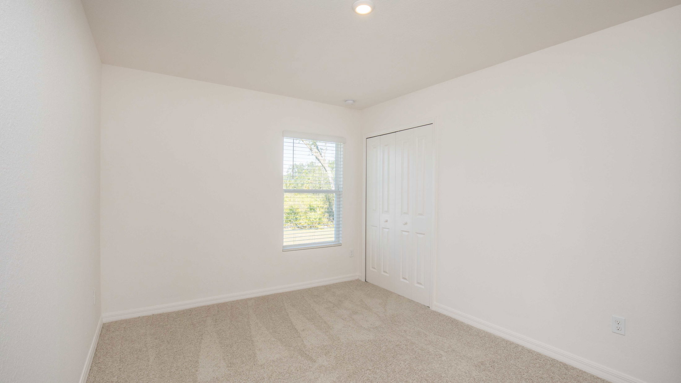 Empty white room with beige carpet, a window with blinds, and a closed closet door under recessed lighting.