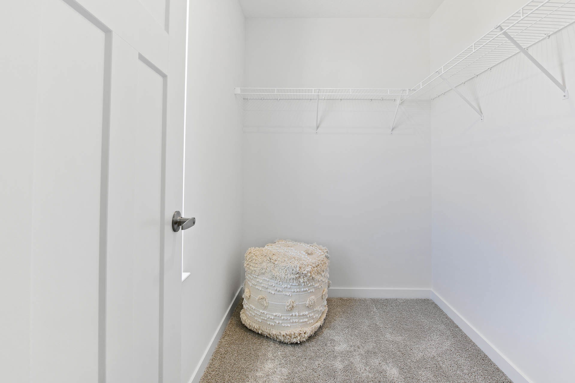 Minimalist empty walk-in closet with white walls, metal wire shelves, and a textured pouf on gray carpet.