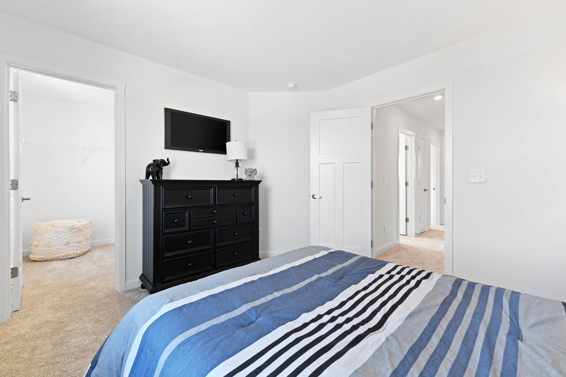 Modern bedroom with a black dresser, flat-screen TV, and striped bedding, leading to a walk-in closet and hallway.