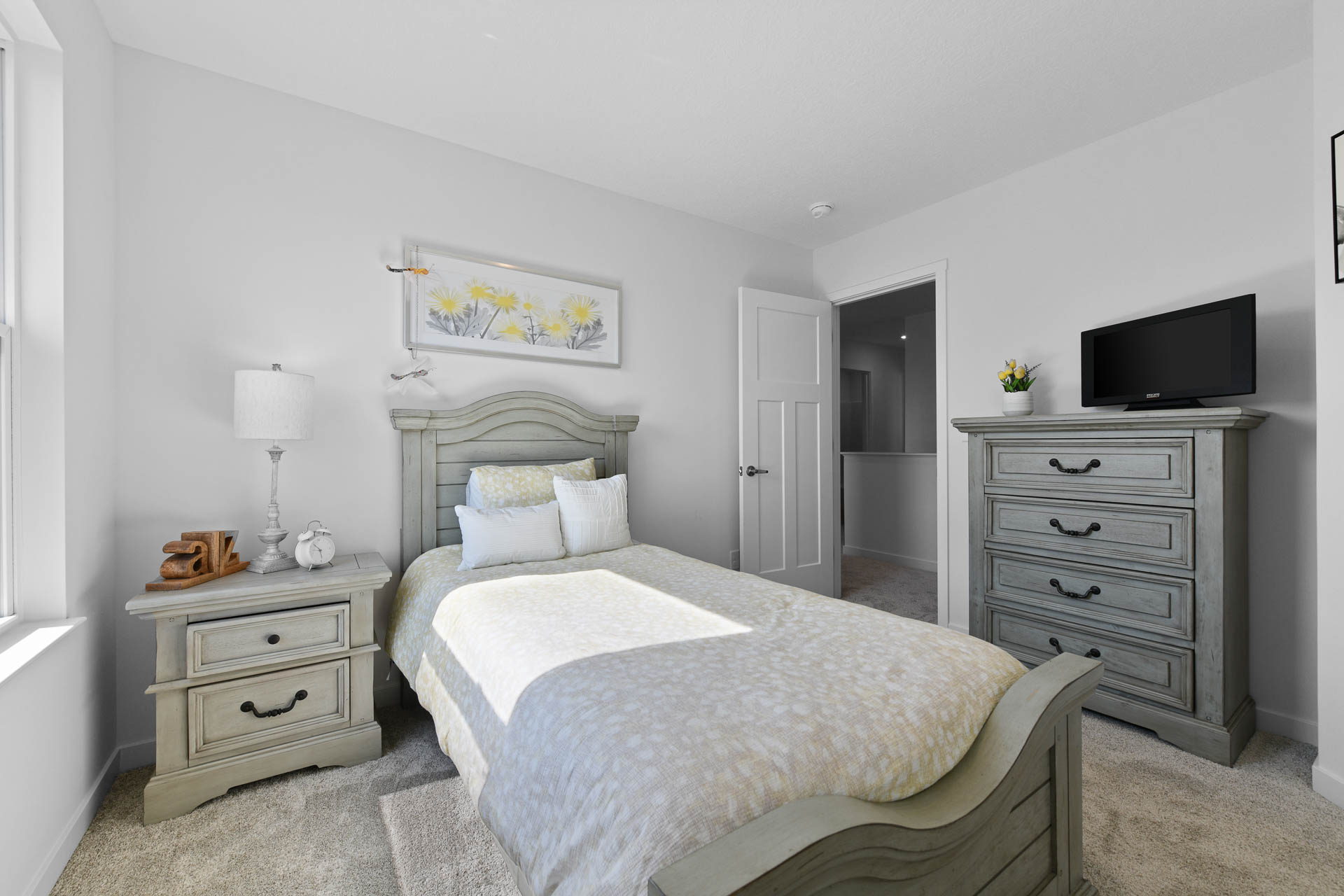 A bright and cozy bedroom featuring a single bed with light bedding, a wooden nightstand and dresser, a wall-mounted TV, and floral artwork.