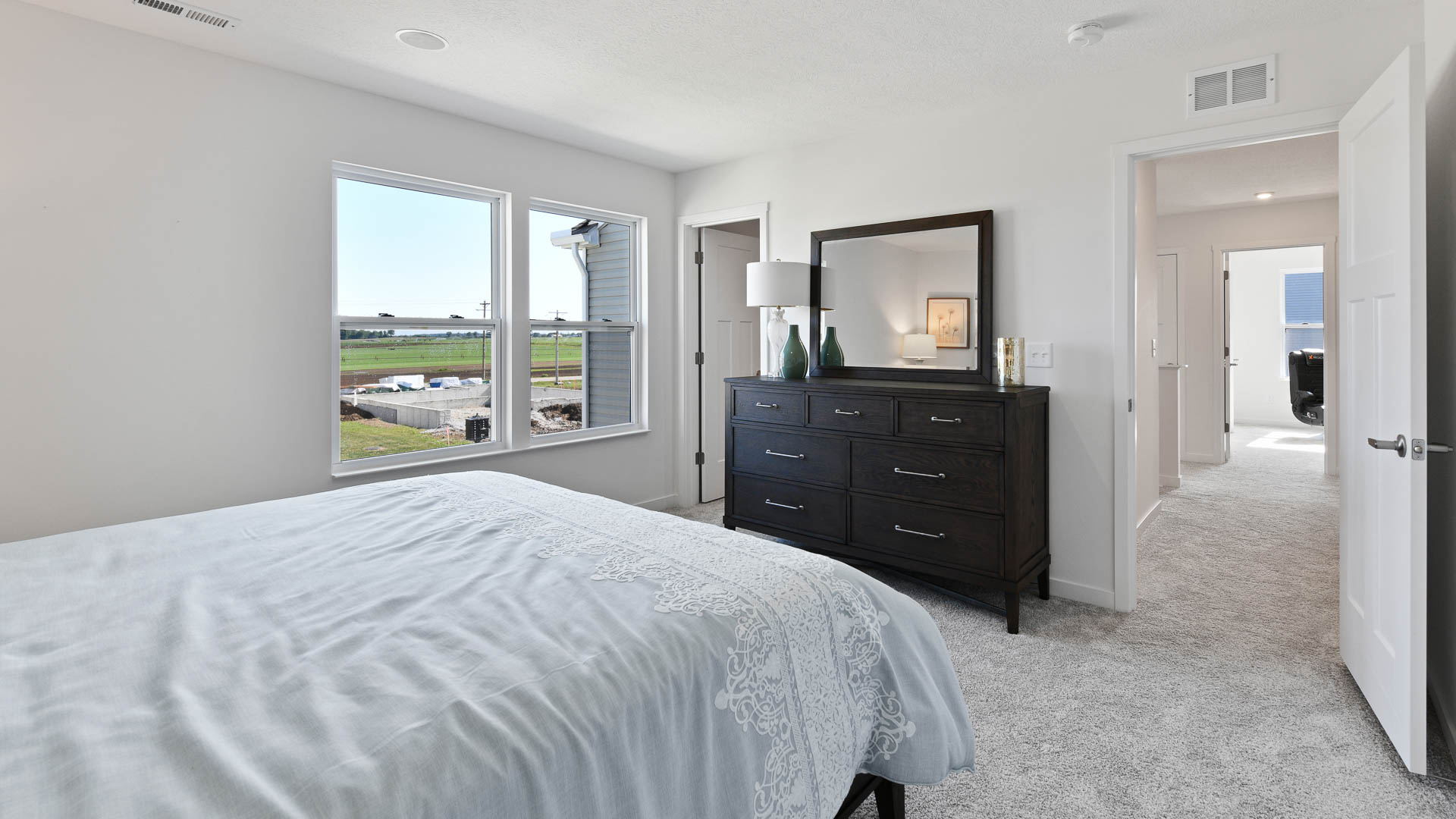 Modern bedroom featuring a large window with a scenic view, a king-sized bed with intricate bedding, and a dark wooden dresser with a mirror and decor.