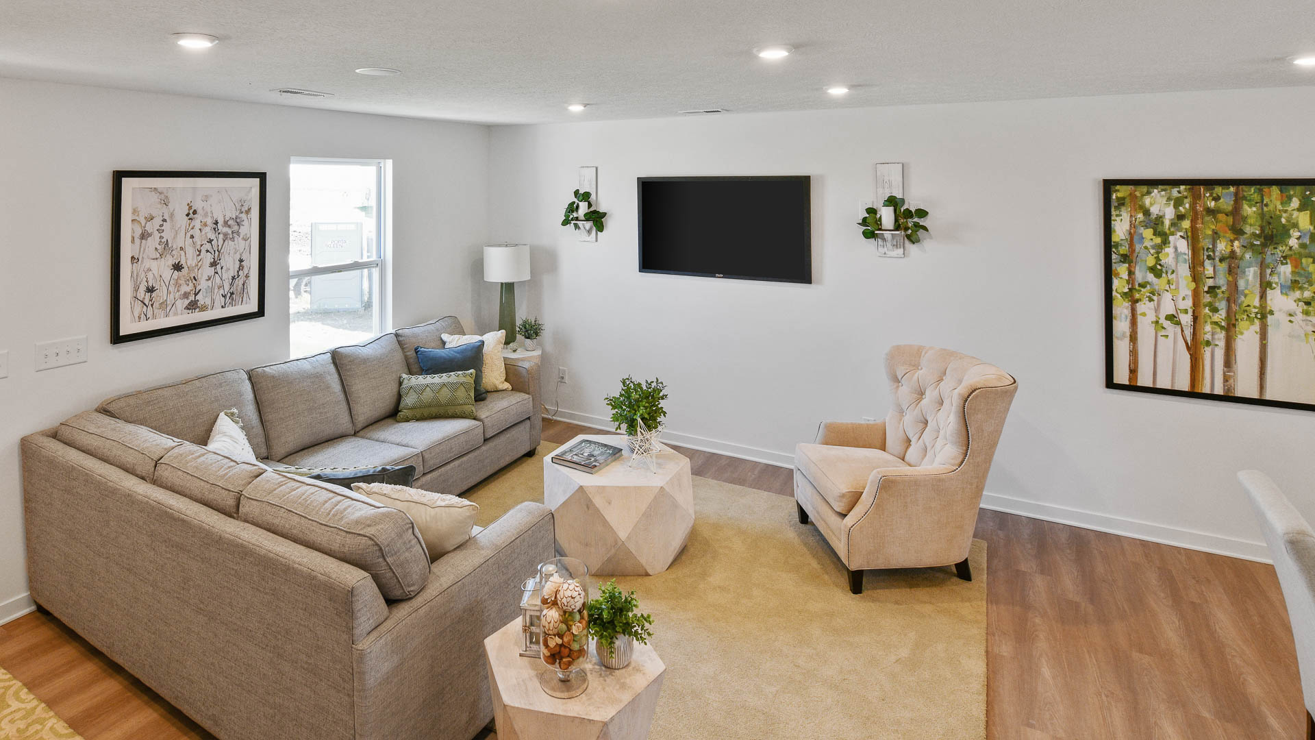 Stylish modern living room with a gray sectional sofa, beige accent chair, mounted flat-screen TV, geometric wooden tables, and framed wall art.