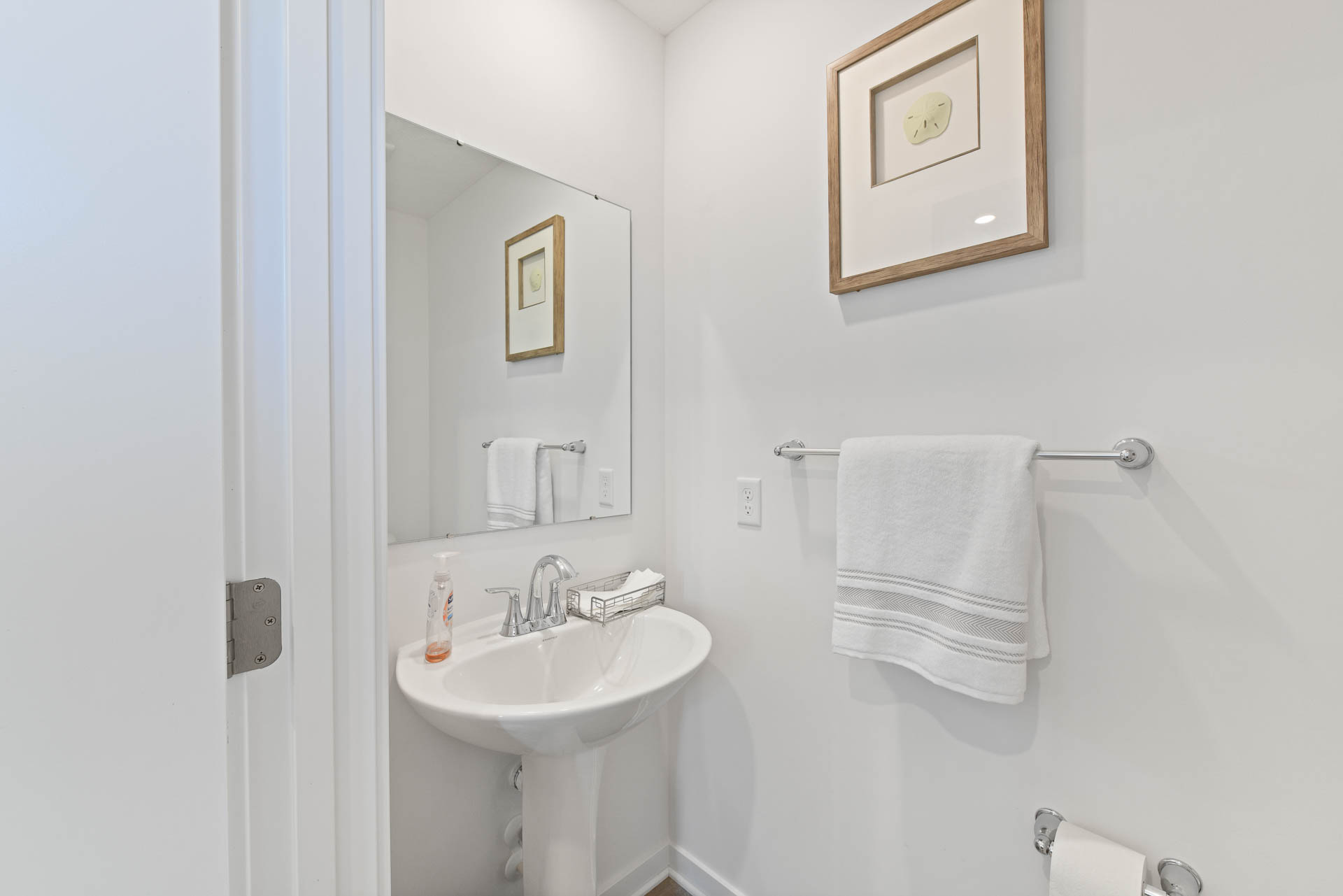 Small modern white bathroom with a pedestal sink, wall mirror, towel rack, and framed wall art.