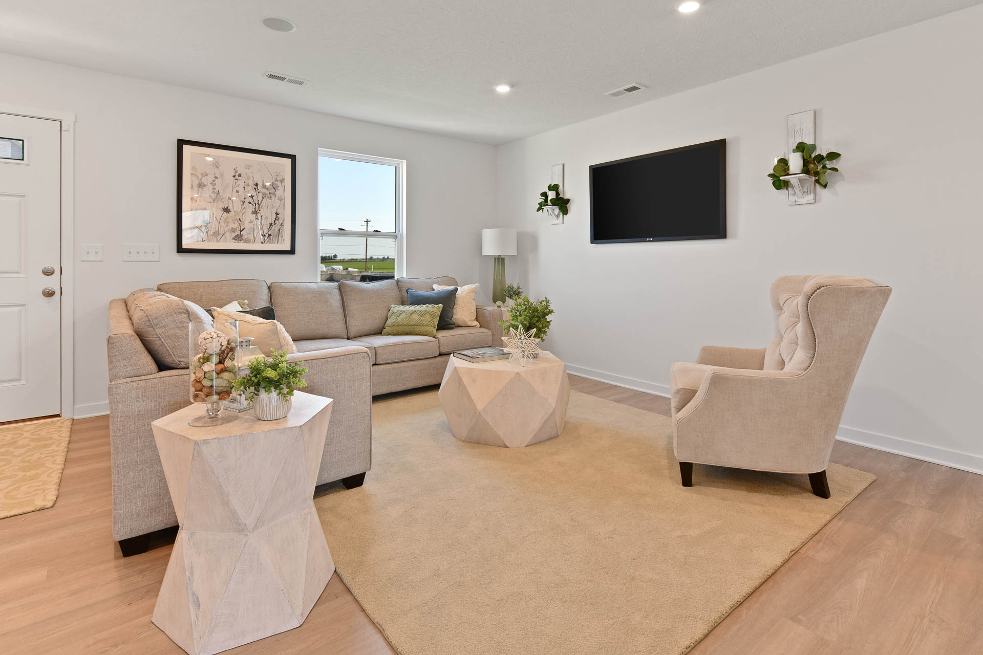 Modern living room with beige sofa and armchair, geometric coffee tables, wall-mounted TV, and floral decor.