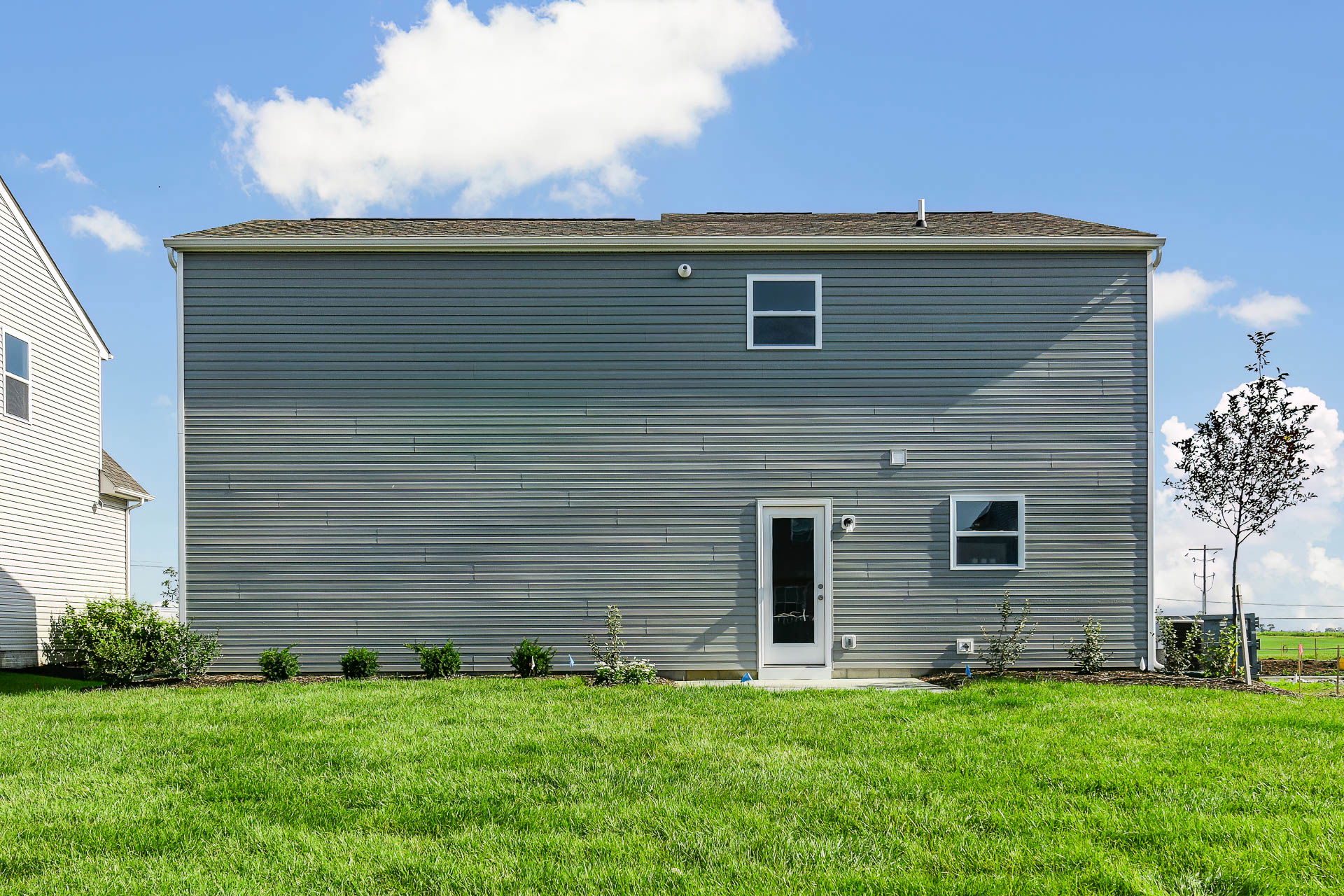 Backyard view of a modern gray two-story house with white trim, featuring a lush green lawn, small shrubs, and blue sky with clouds.