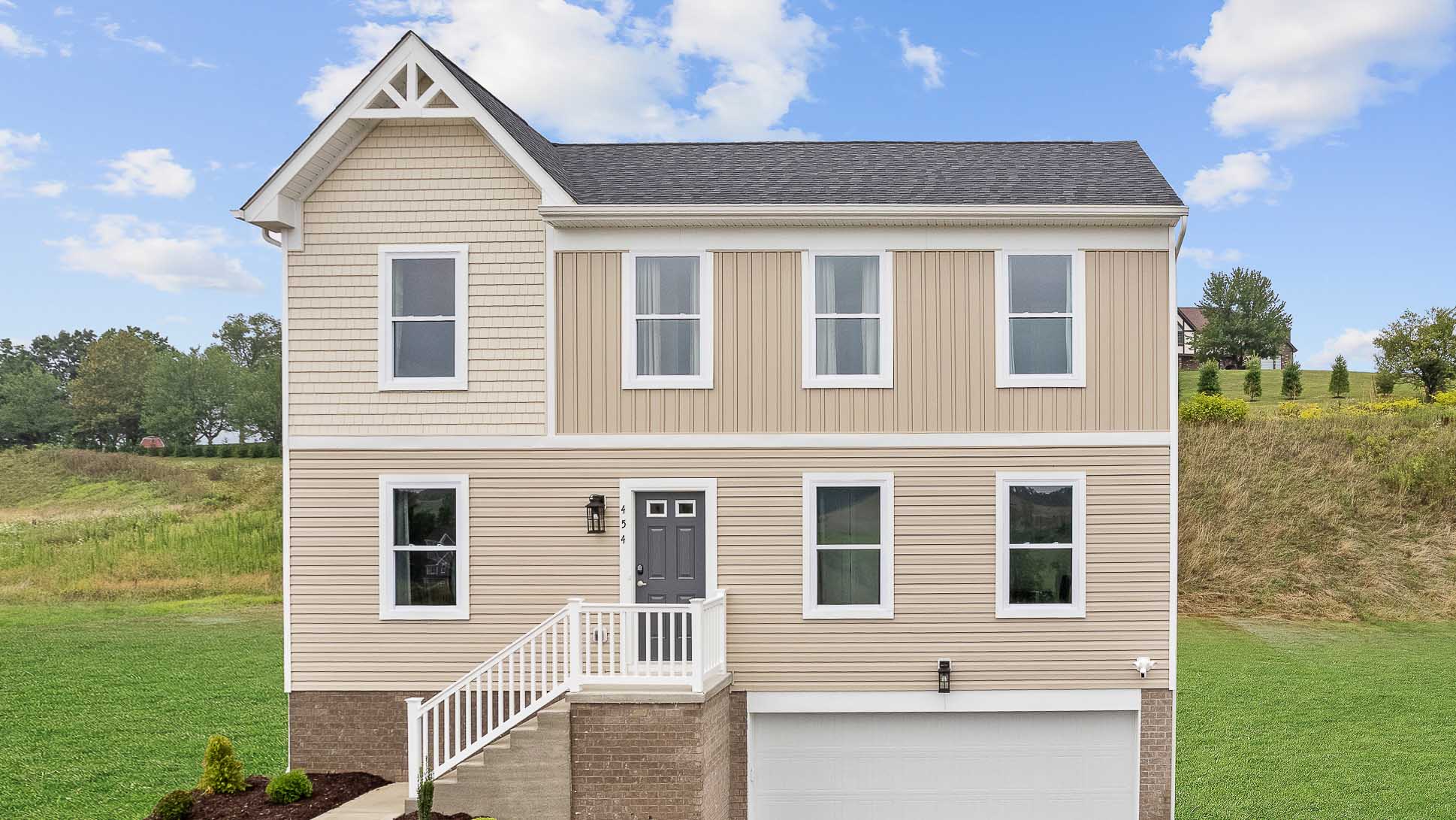 Two-story beige house with gray door, front porch, and a green lawn, under a blue sky with clouds.