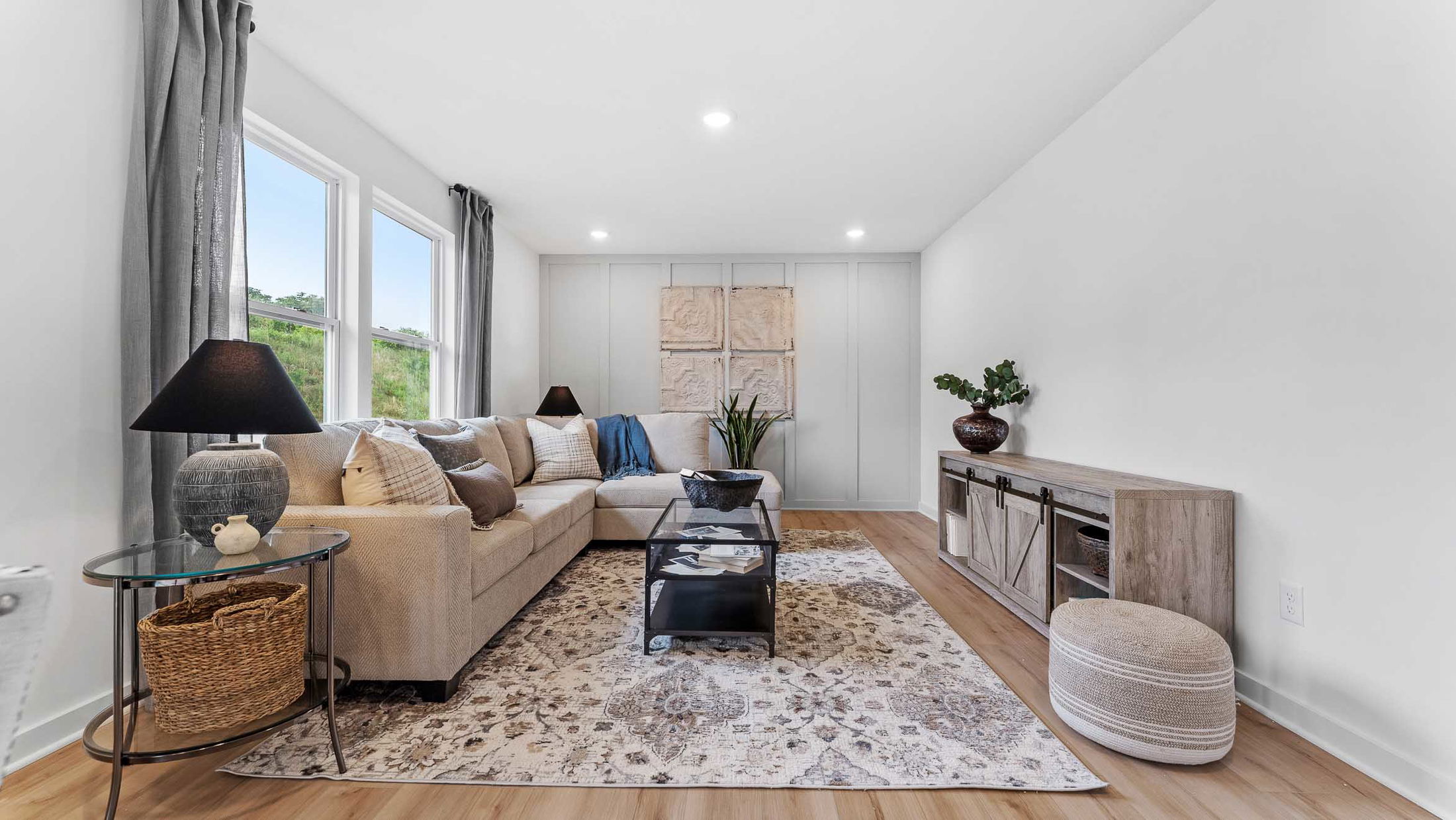 A modern living room with neutral decor features a beige sectional sofa, black table lamps, glass coffee table, and a rustic wooden console table.