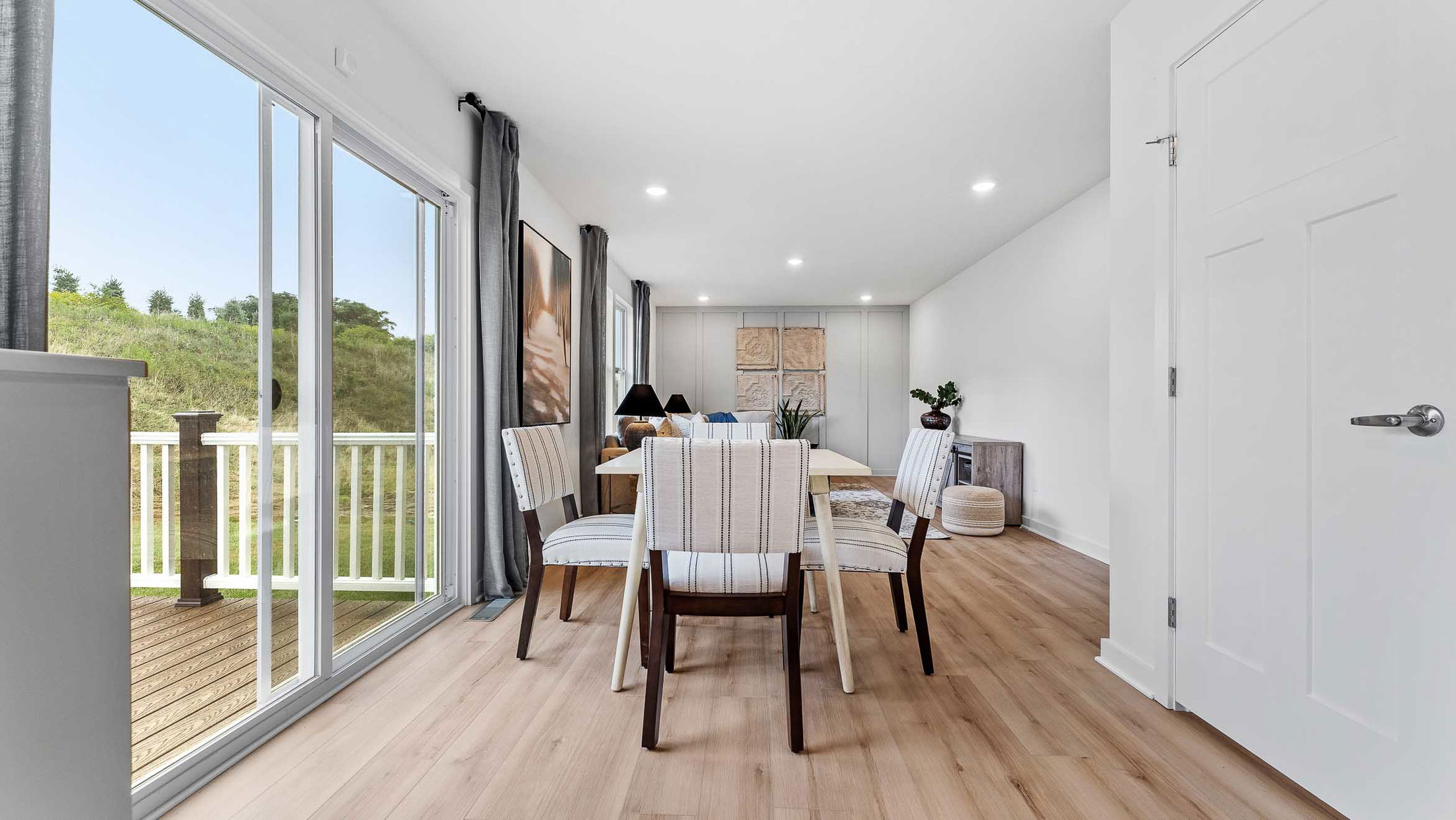 Bright modern dining area with striped chairs, wooden flooring, and large sliding glass doors opening to a deck and green landscape.