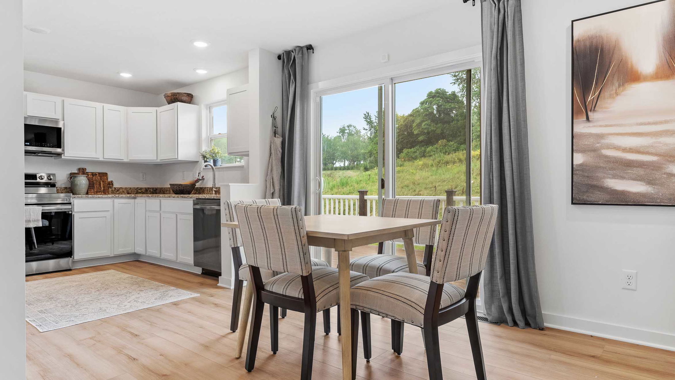 Bright, modern kitchen and dining area with white cabinets, wooden flooring, and large windows offering a scenic view of greenery.