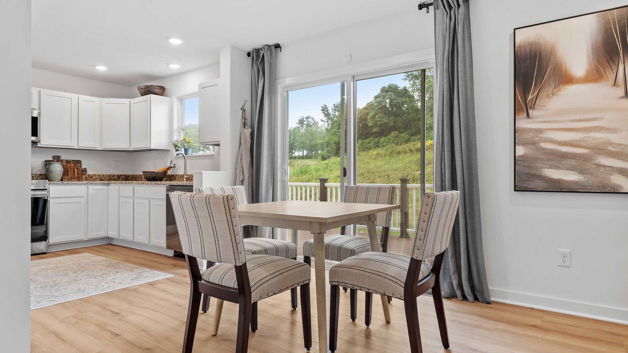 Modern kitchen and dining area with white cabinets, wooden floors, and a view of lush greenery through large sliding glass doors.