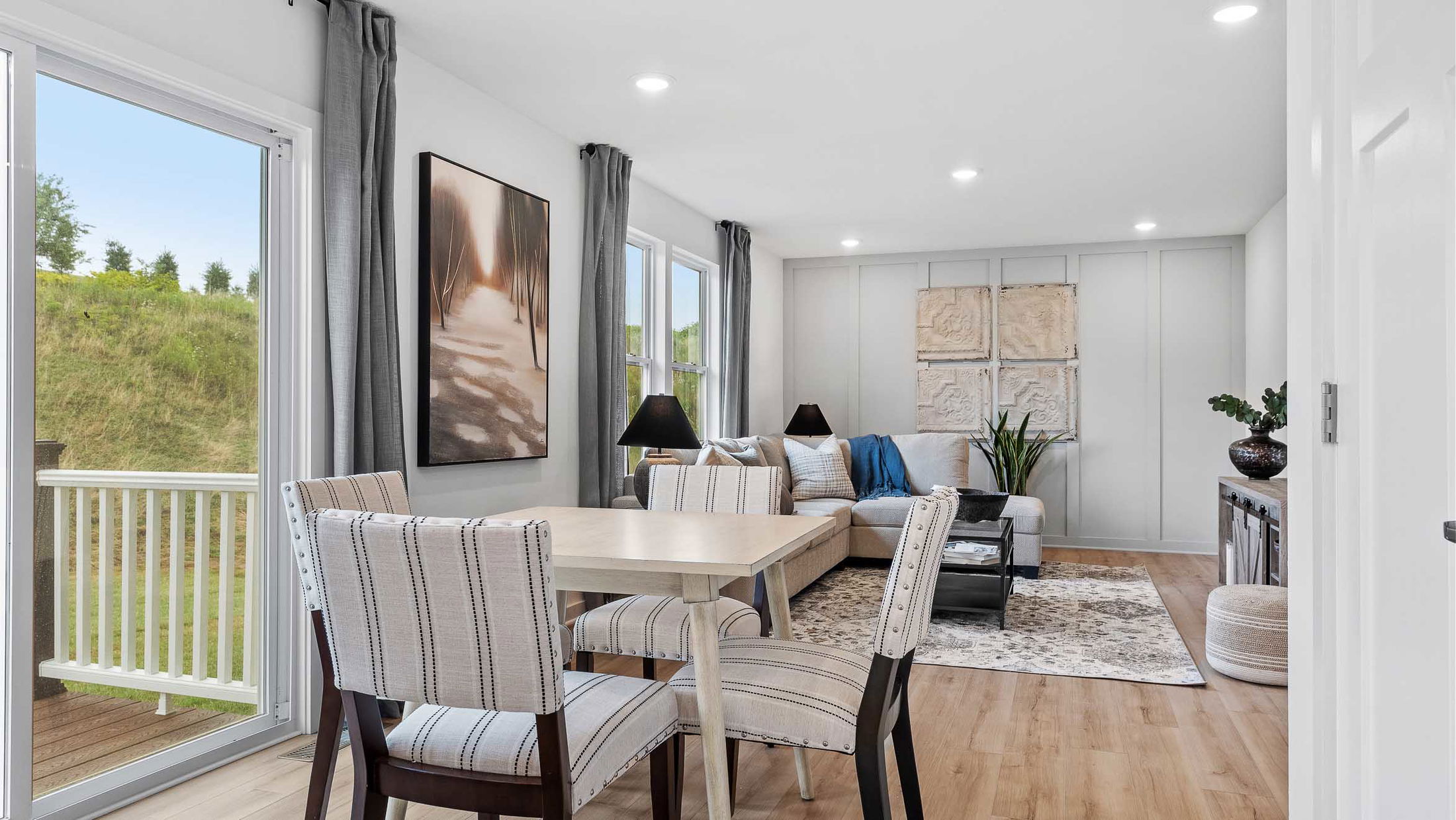 Modern open-concept living and dining room with stylish decor, featuring a light wood floor, elegant grey curtains, and scenic outdoor view.