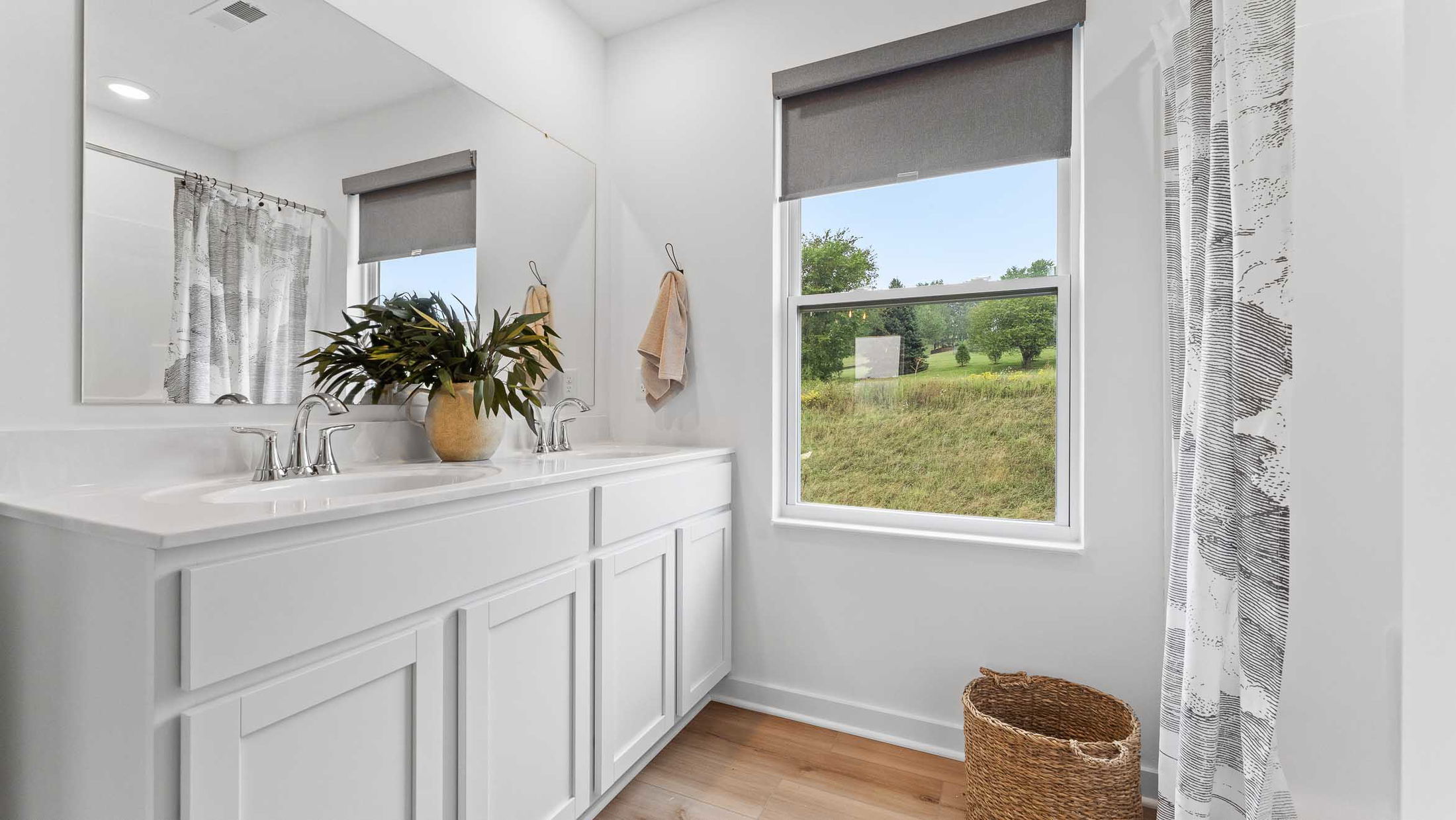 Bright, modern bathroom with white cabinetry, large mirror, potted plant, wicker basket, and window overlooking a lush green landscape.