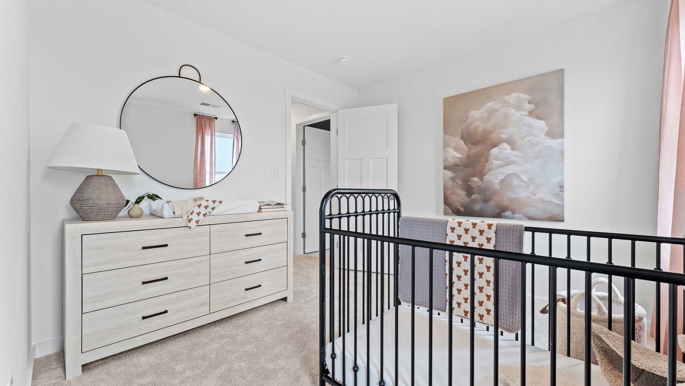 Modern baby nursery with a dark crib, white dresser with a round mirror, and soft cloud artwork.