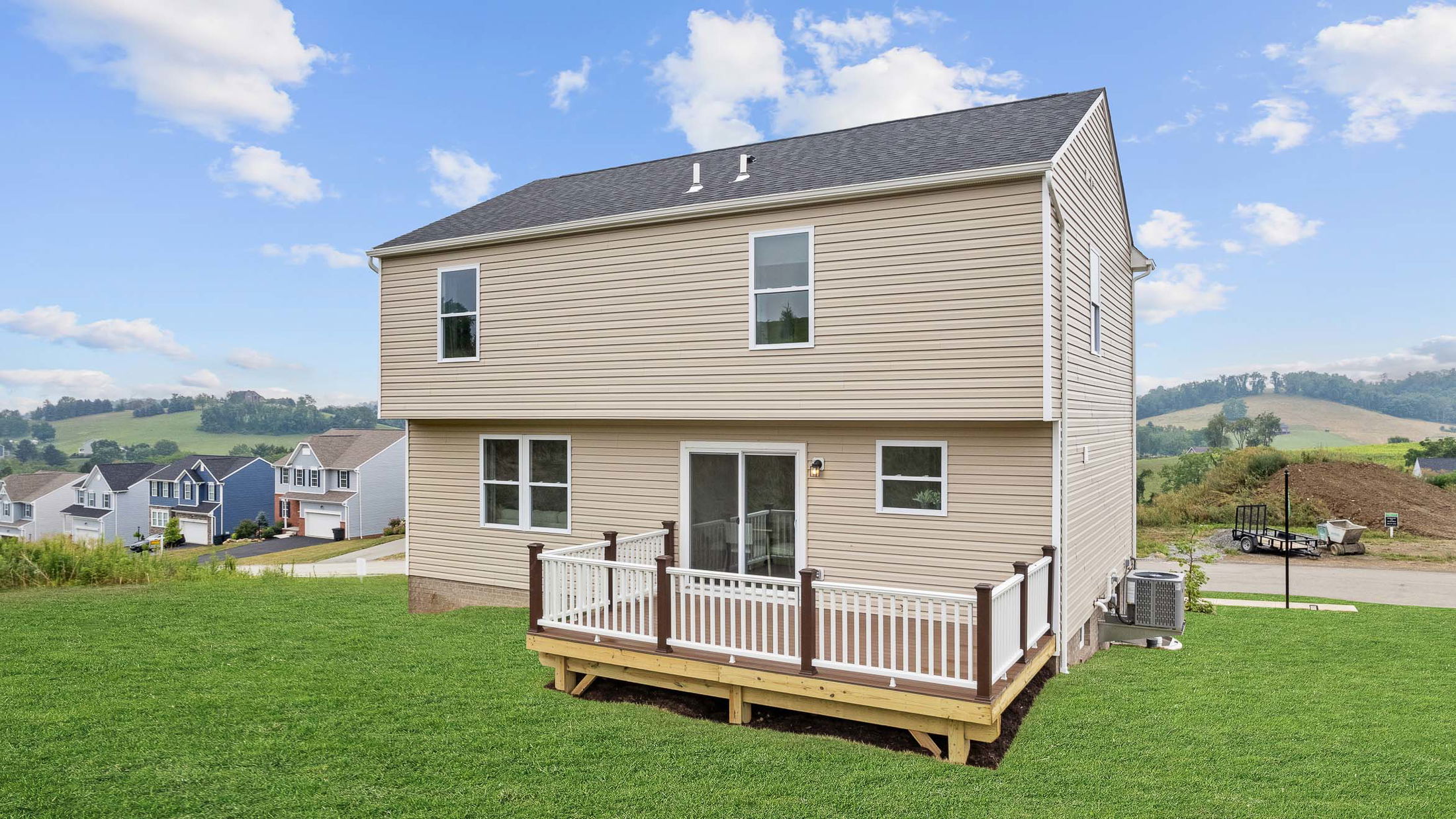 Modern two-story beige house with a wooden deck, surrounded by green grass and hills.