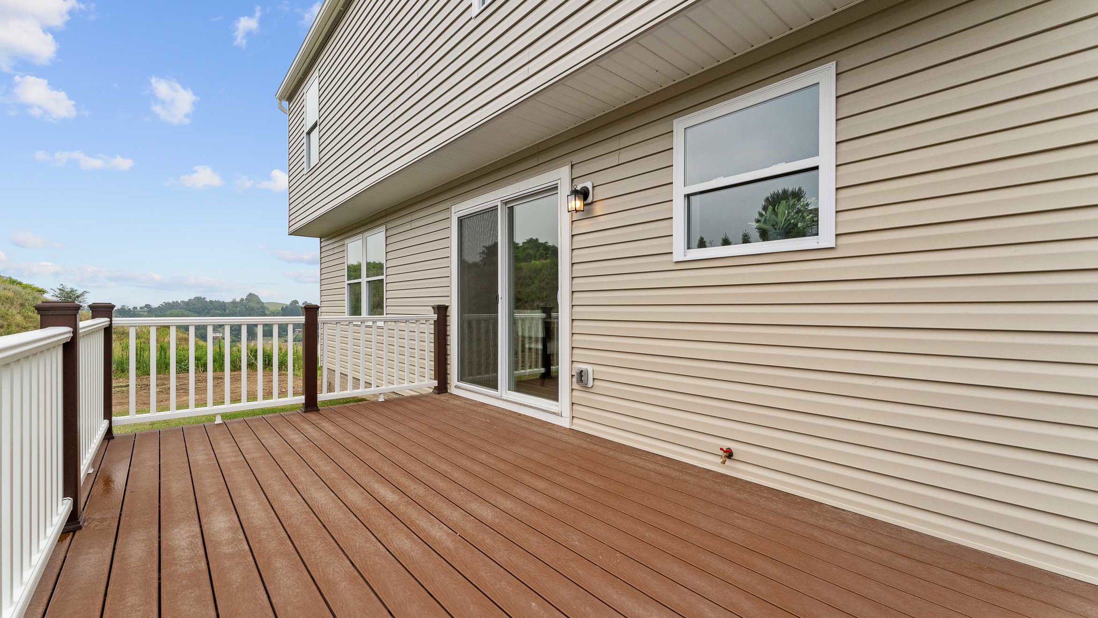 Spacious wooden deck with white railing attached to a beige vinyl-sided house overlooking a lush green landscape.