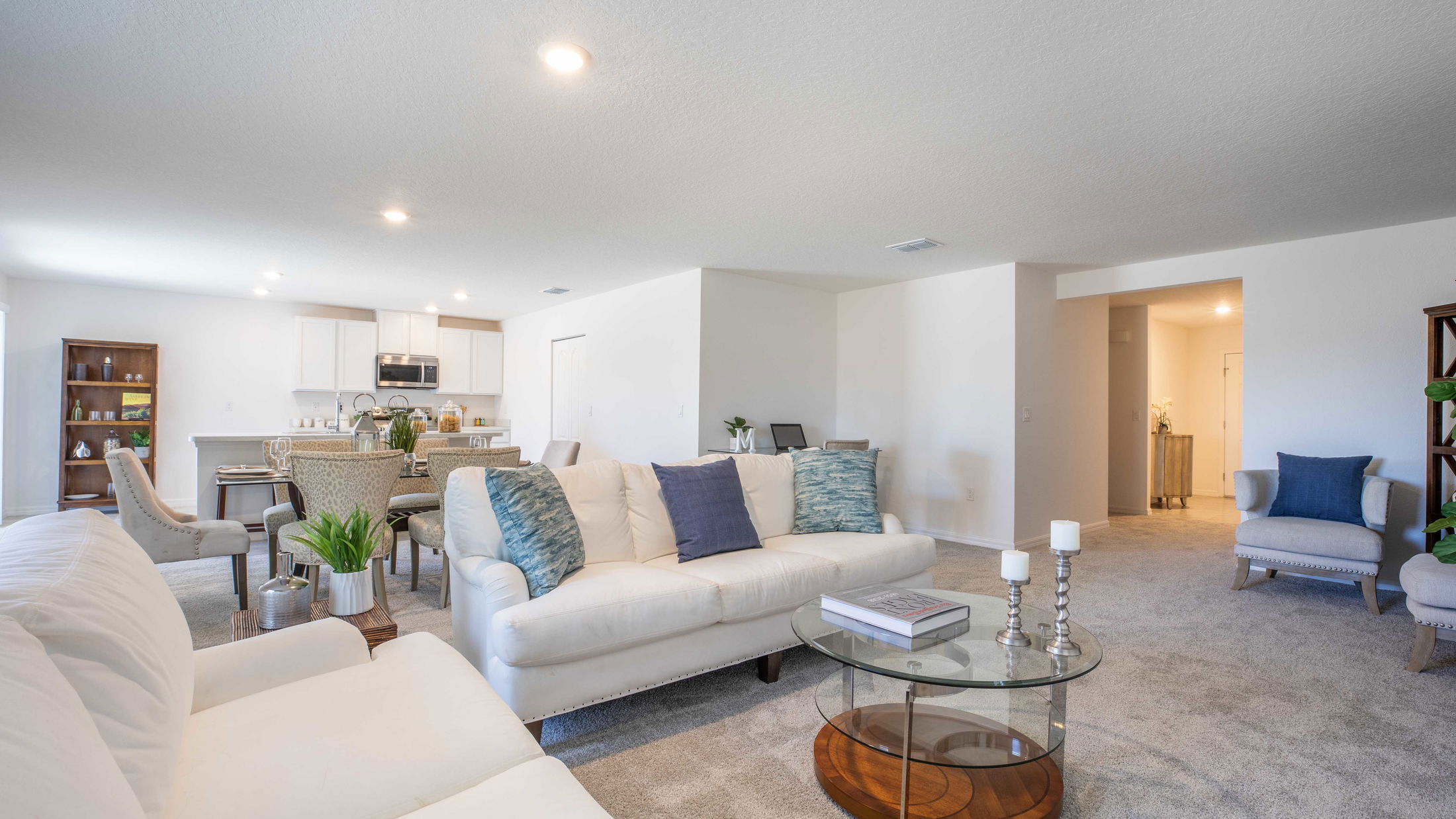 Modern open-concept living room with white sofas, a glass coffee table, and a view of the dining area and kitchen.