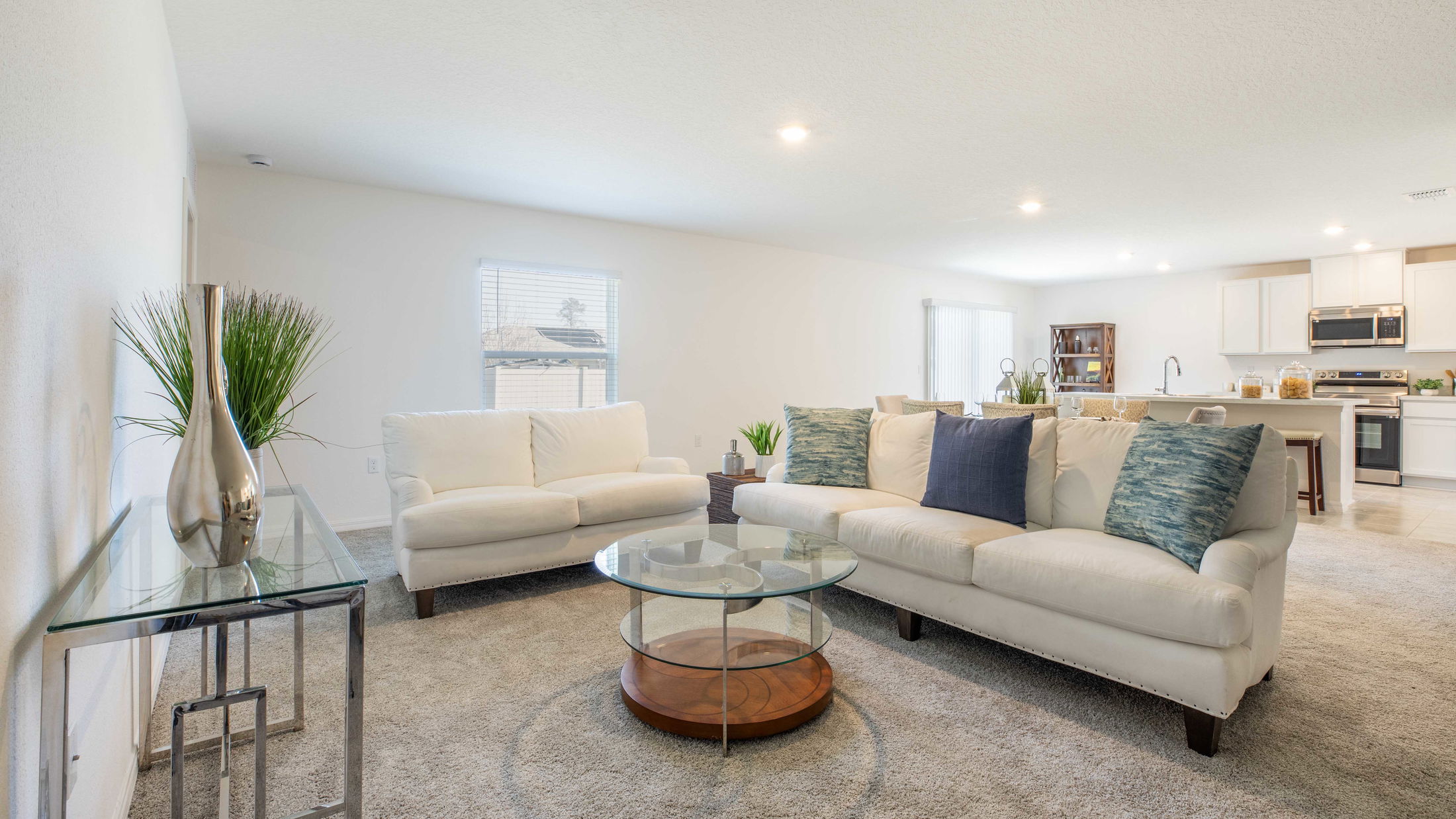 Modern living room with white sofas, a glass coffee table, decorative vases, and open kitchen layout.