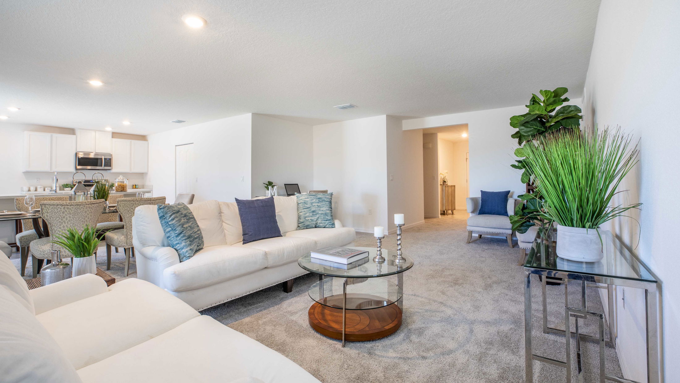 Spacious and elegant modern living room and kitchen with white sofas, decorative cushions, green plants, and sleek furniture.