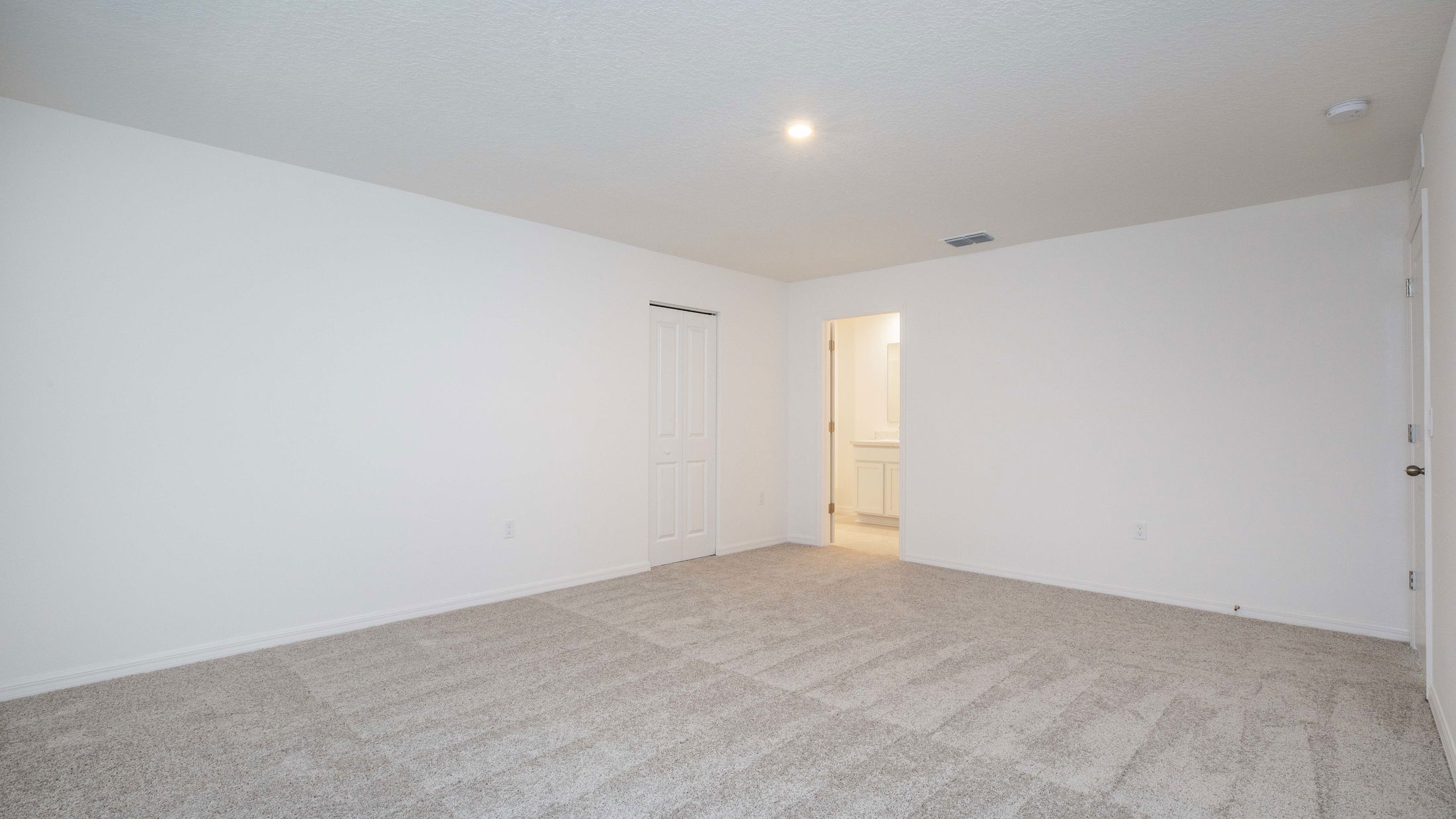 Empty neutral-colored room with beige carpet and white walls leading into a bathroom, ideal for real estate listings.