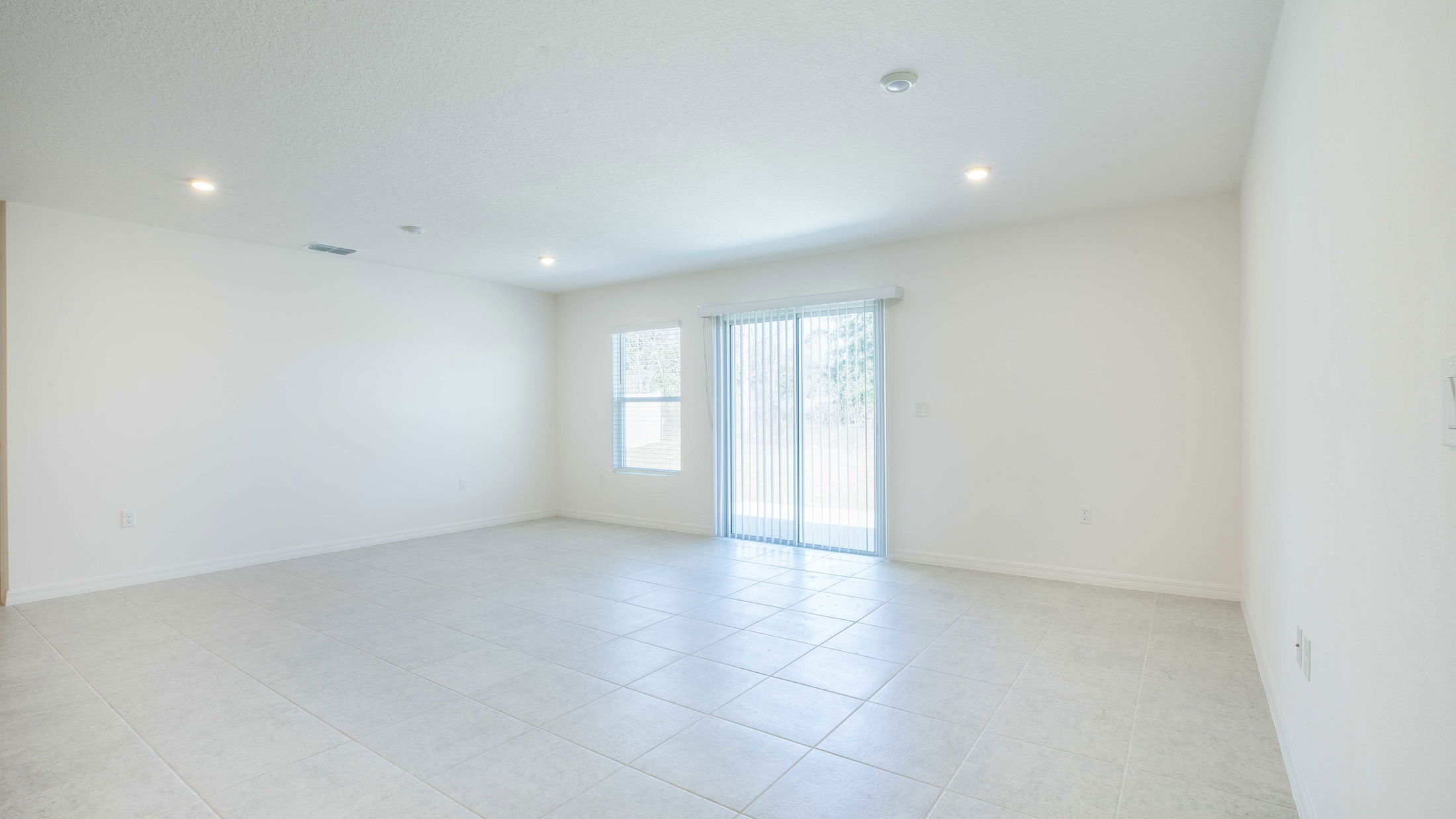 Bright, unfurnished living room with large tile flooring, white walls, and sliding glass doors allowing natural light.
