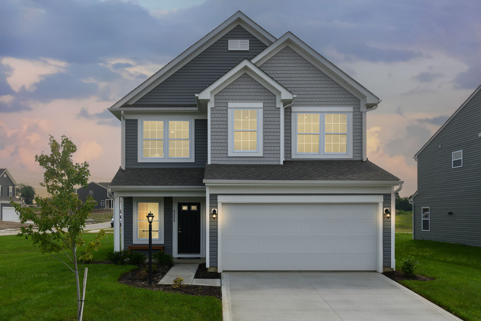 Modern two-story gray suburban house with lit windows, a two-car garage, and a well-maintained lawn during sunset.