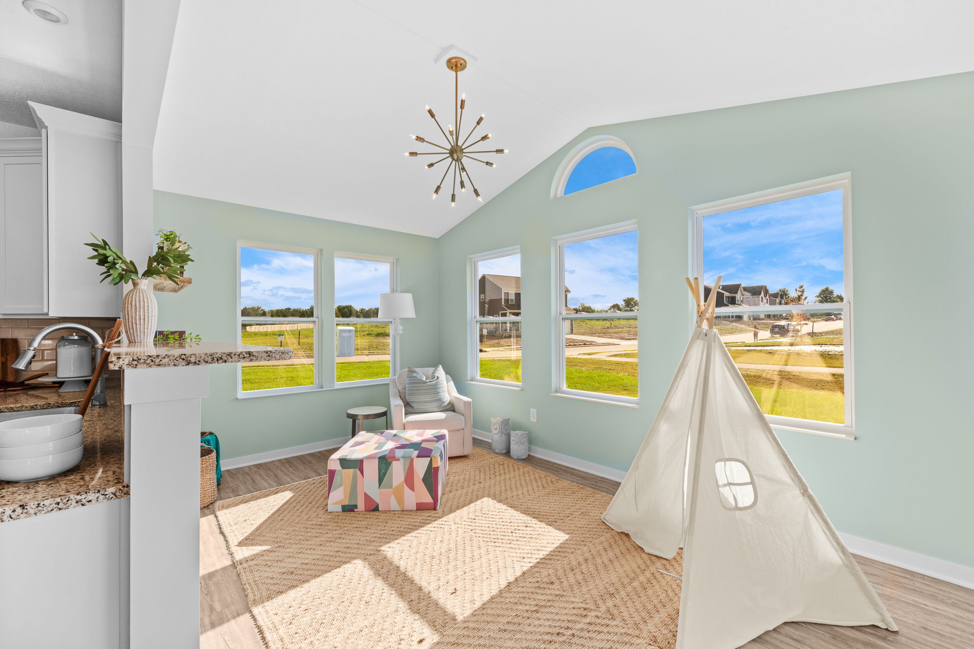 Bright sunroom with large windows, modern lighting, colorful ottoman, and a children's play tent, overlooking a scenic suburban neighborhood.