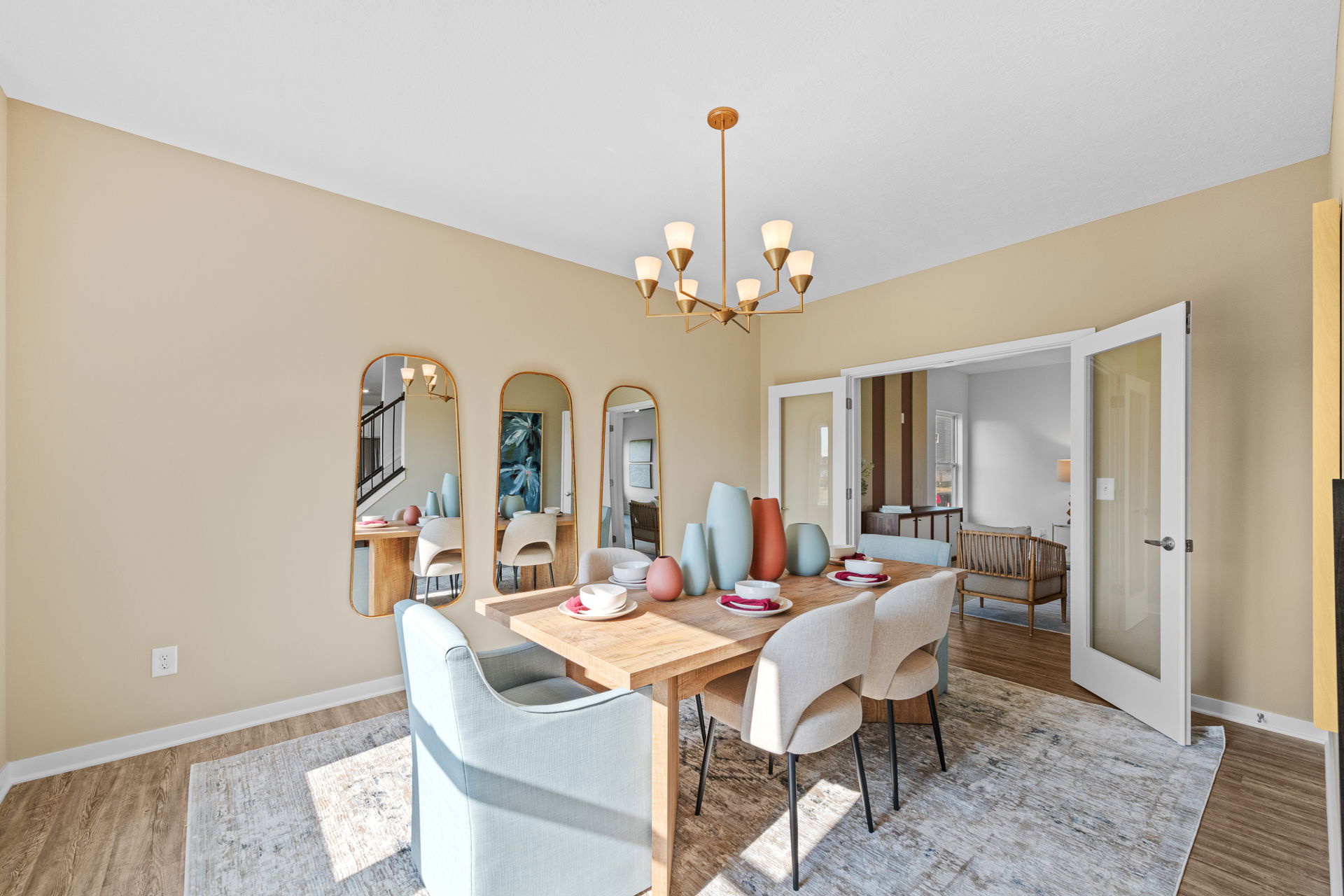 Modern dining room with wooden table, pastel chairs, decorative vases, and wall mirrors under a stylish chandelier.