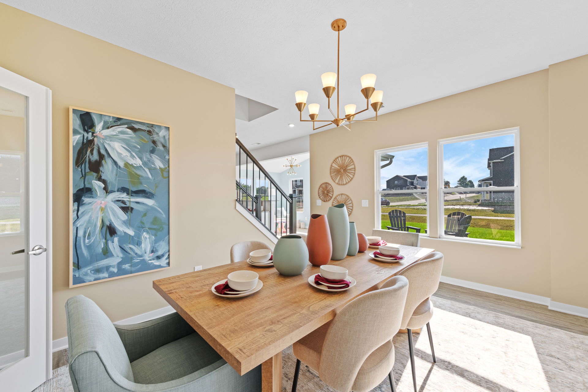 Bright, modern dining room featuring a wood table set with colorful vases, surrounded by upholstered chairs and accented by a floral painting and contemporary chandelier.