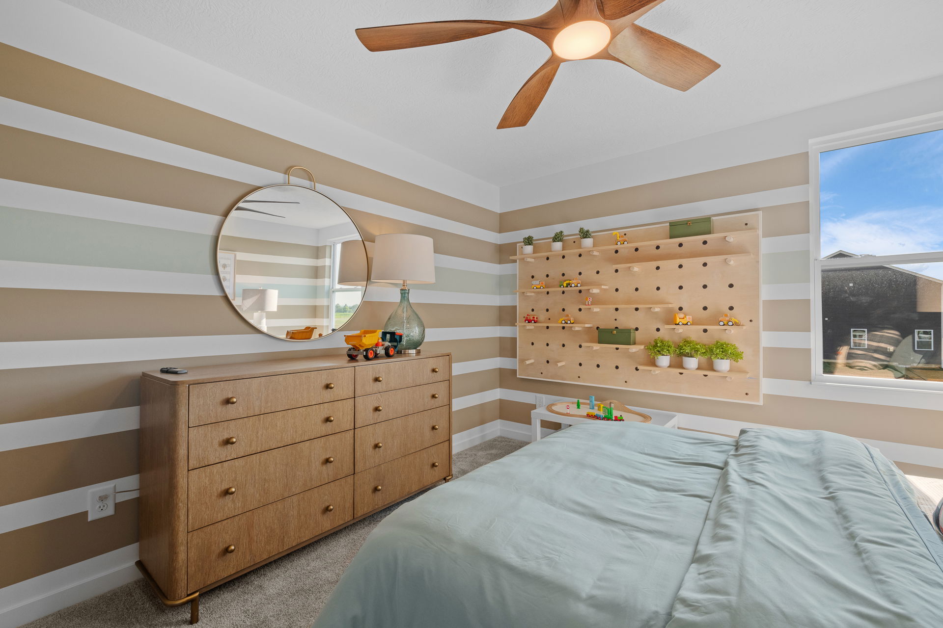 Modern bedroom with striped walls, a round mirror above a wooden dresser, and a pegboard display with plants and toys.