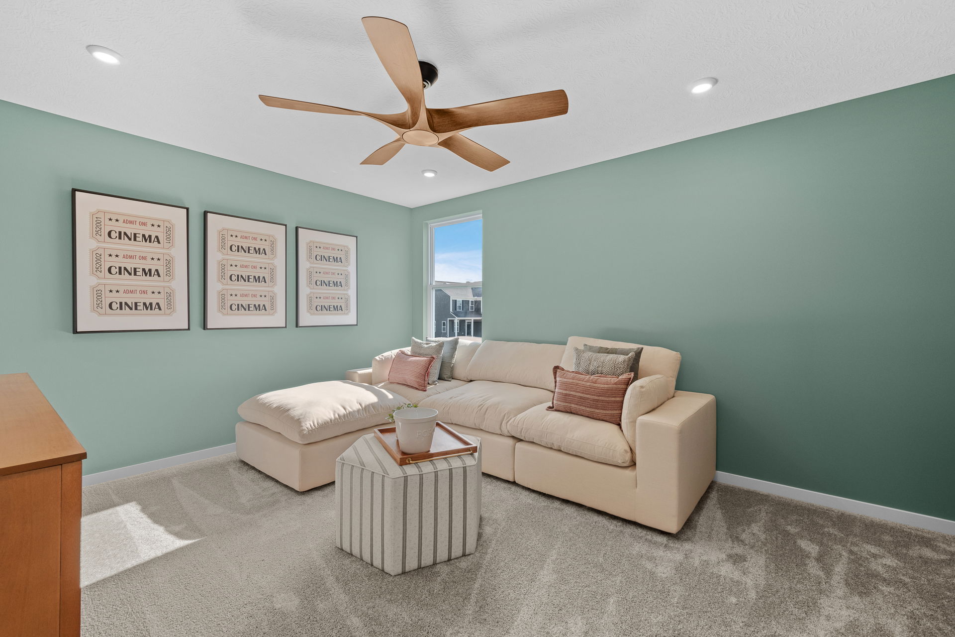 Modern living room with a beige sectional sofa, mint green walls featuring cinema ticket art, large window, and wooden ceiling fan.