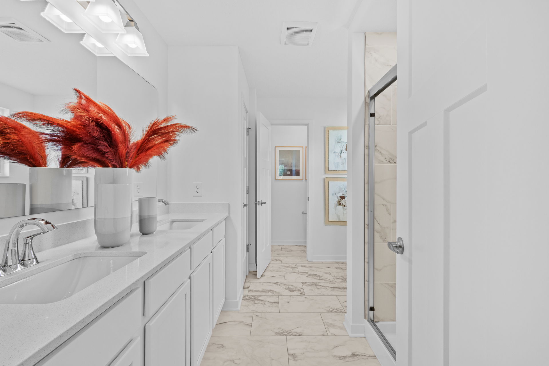 Modern white bathroom interior featuring dual sinks, marble tile flooring, a glass shower, and a vibrant red floral arrangement.