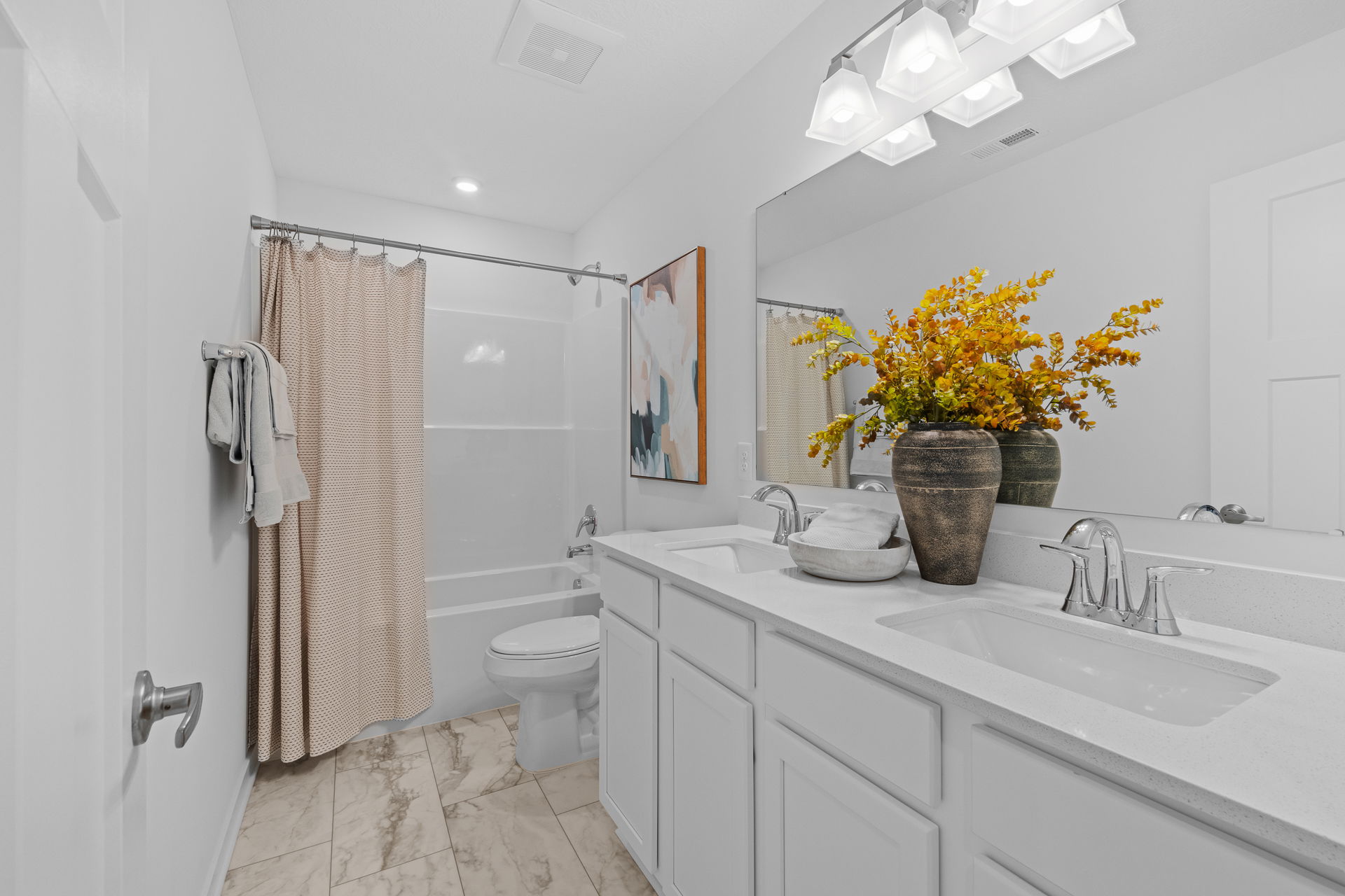 Modern bathroom with white fixtures, double sink, large mirror, decorative vase with yellow flowers, and a shower with a beige curtain.
