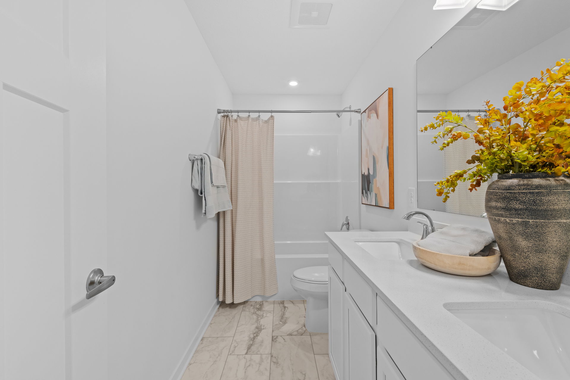 Modern bathroom with white cabinets, a large mirror, marble tile flooring, a shower with a beige curtain, and decorative flowers on the countertop.