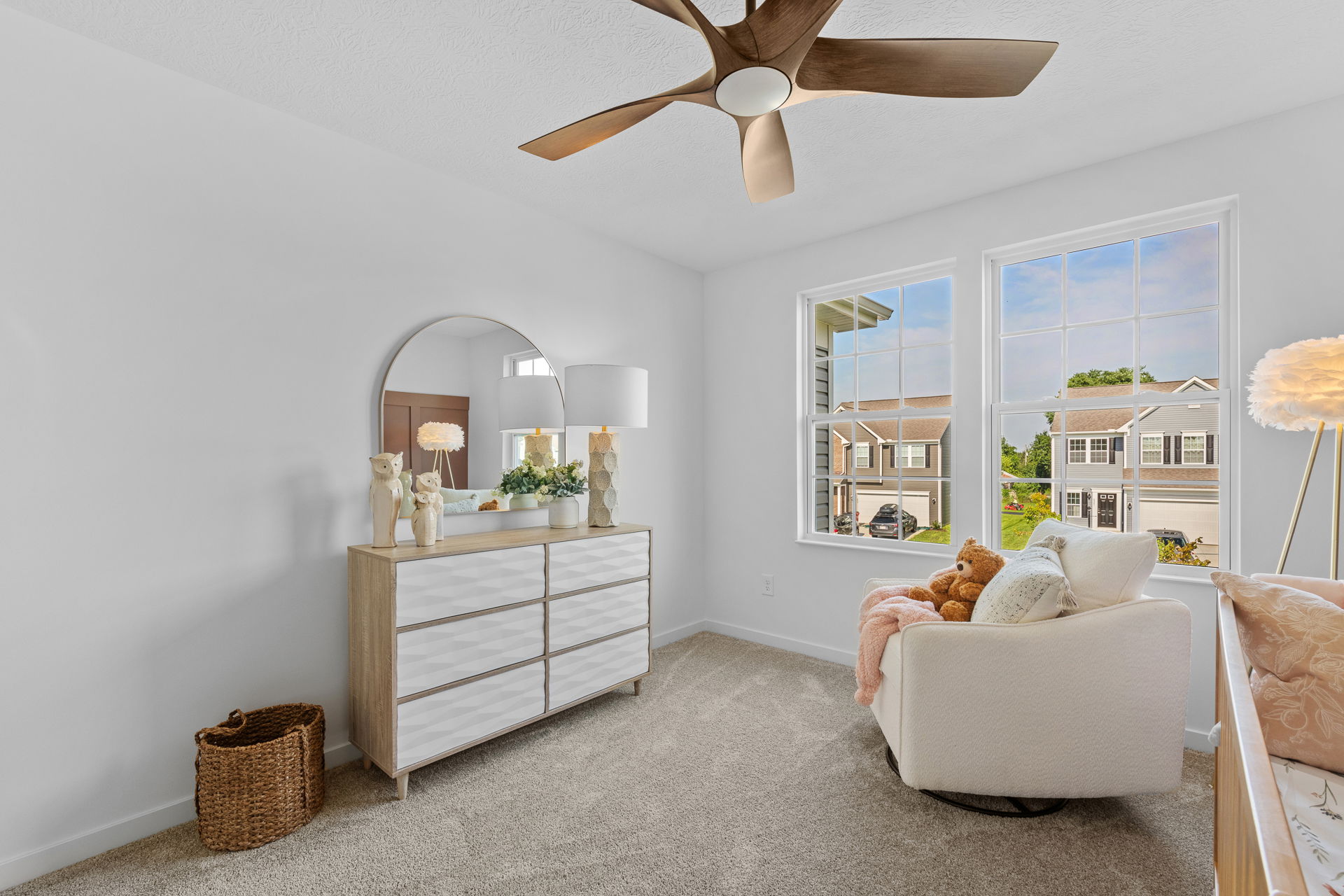 Modern, cozy bedroom with a wooden ceiling fan, dresser with decorative items, and a plush chair by large windows overlooking a suburban street.