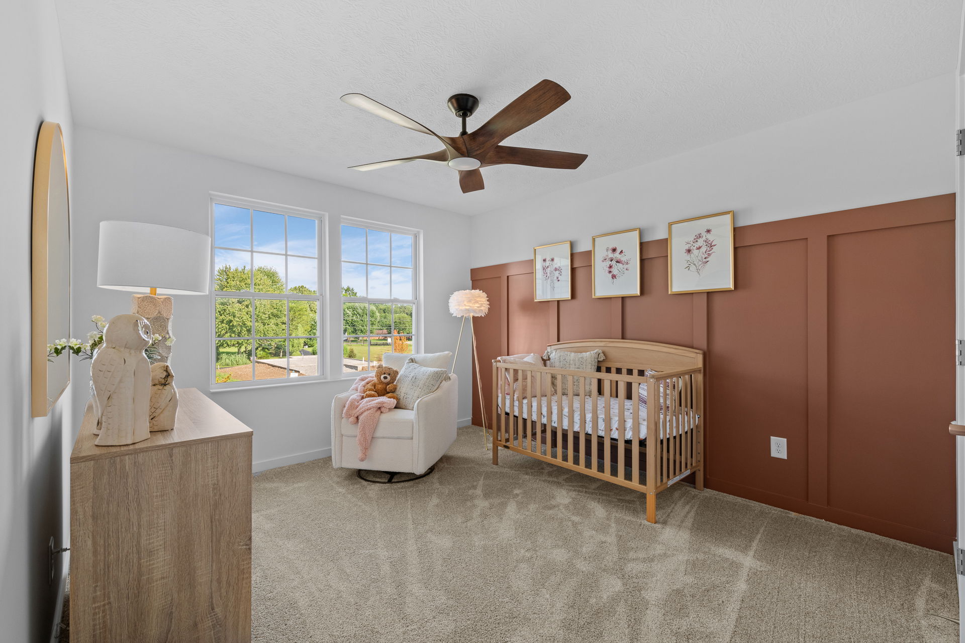 Modern nursery room with a wooden crib, cozy armchair, ceiling fan, and decorative artwork on the wall, featuring large windows overlooking a lush garden.