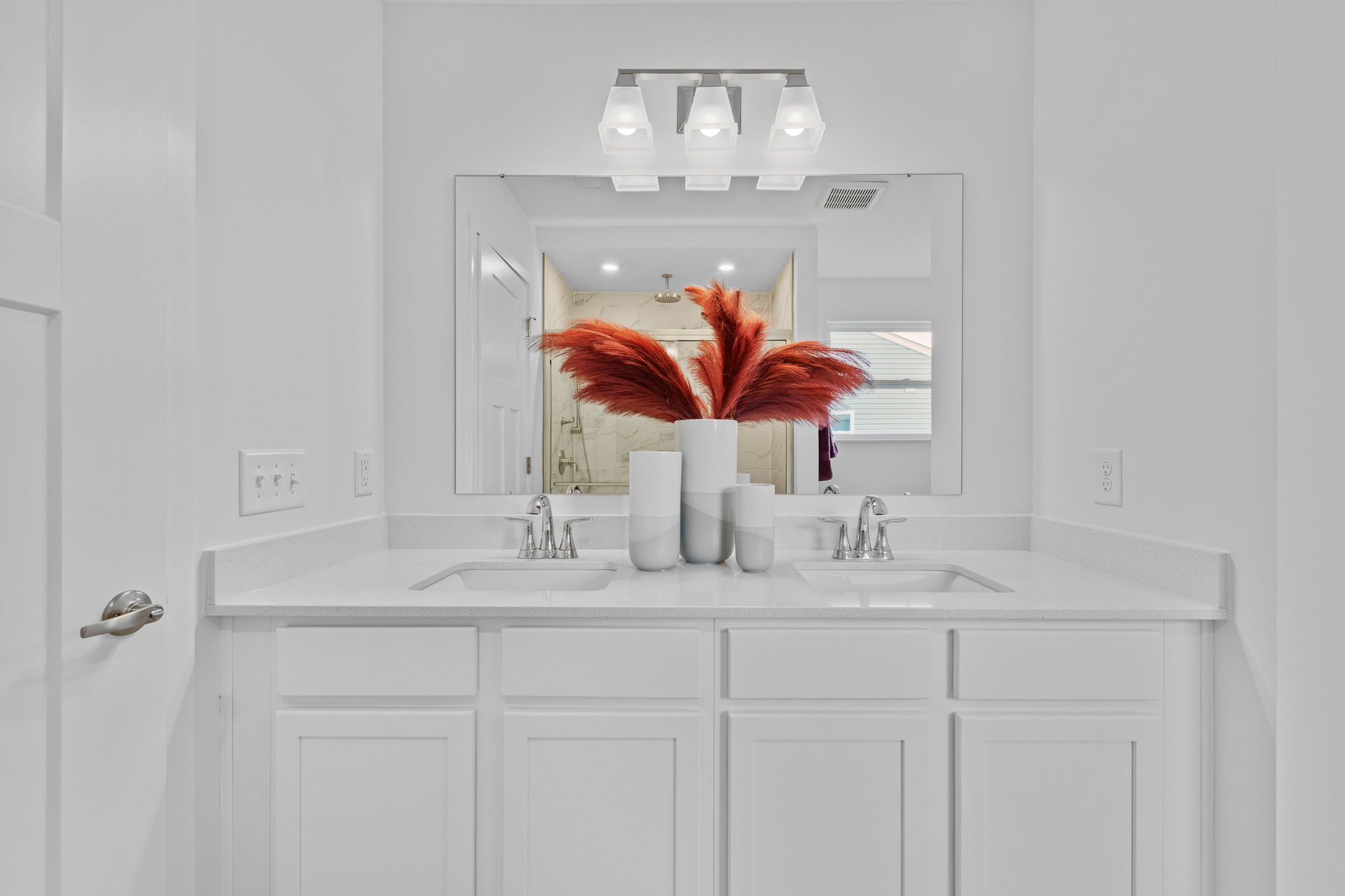 Modern white bathroom with double sinks, large mirror, and red decorative pampas grass in vases for stylish decor.