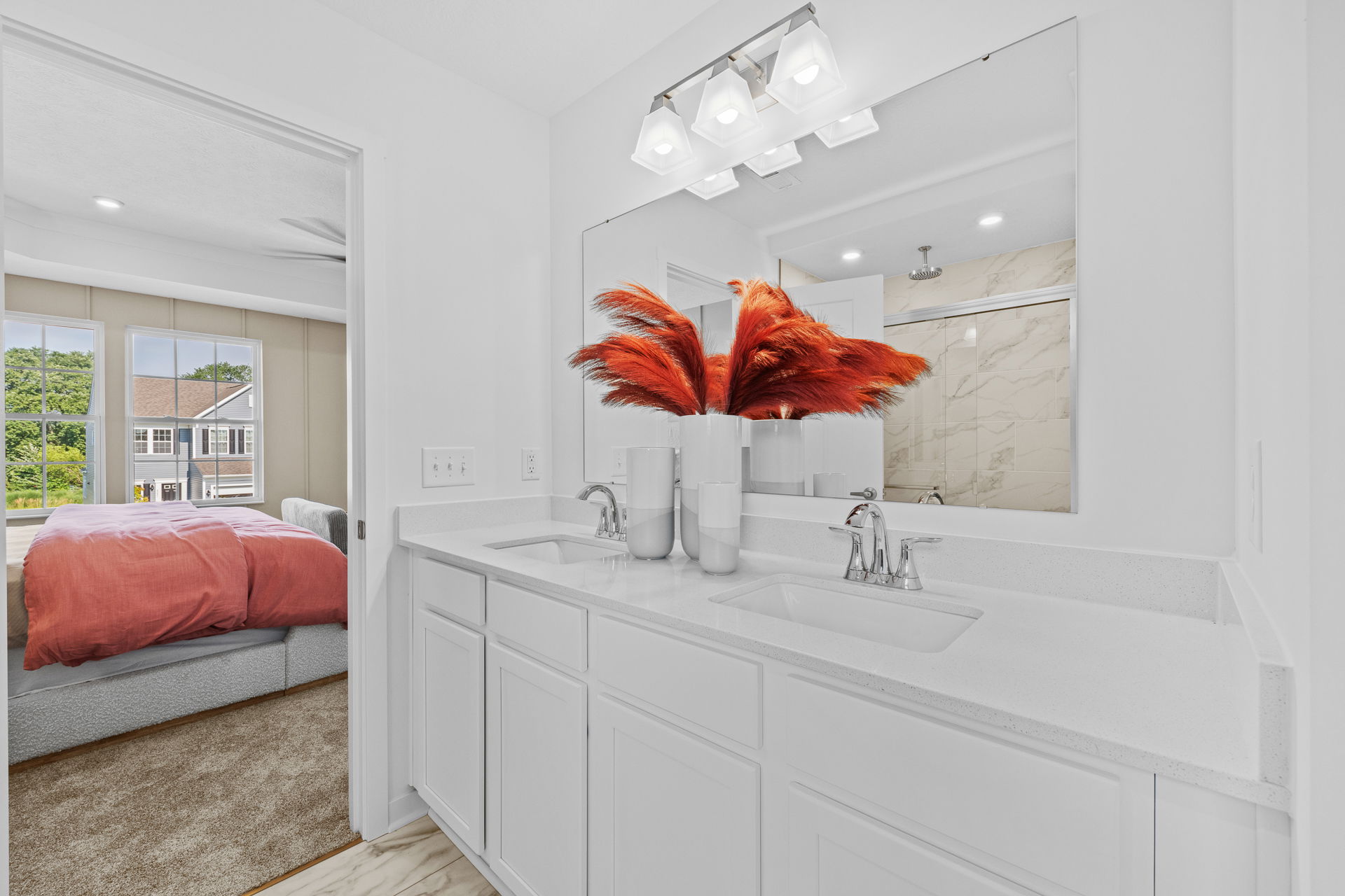 Modern bathroom with white double vanity, large mirror, and vibrant red decorative plants, adjacent to a bedroom with red bedding and natural light.