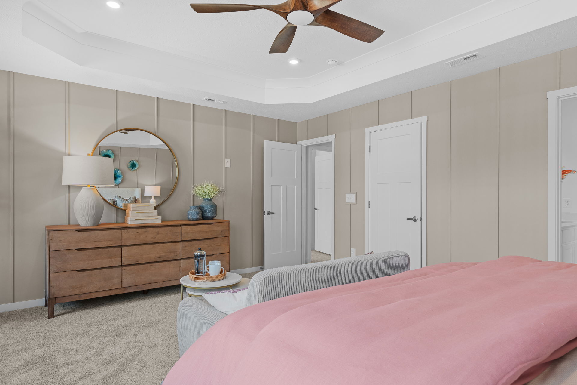 Modern bedroom with beige walls, a wooden dresser, round mirror, and pink bedspread, featuring stylish decor and a ceiling fan.