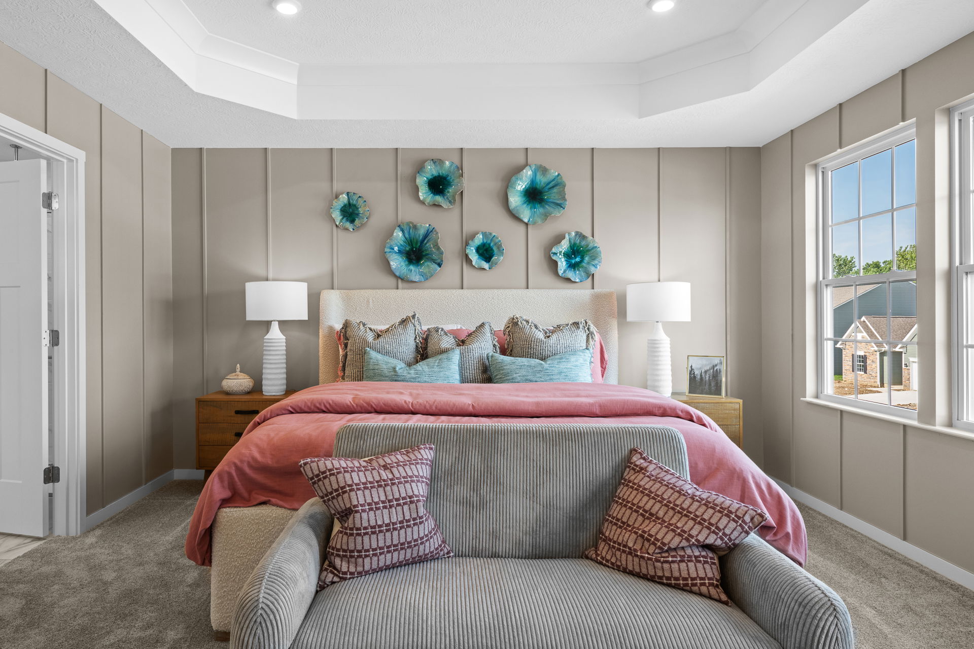 Elegant bedroom interior featuring a cozy bed with pink and blue pillows, decorative wall art, stylish nightstands with lamps, and large windows.