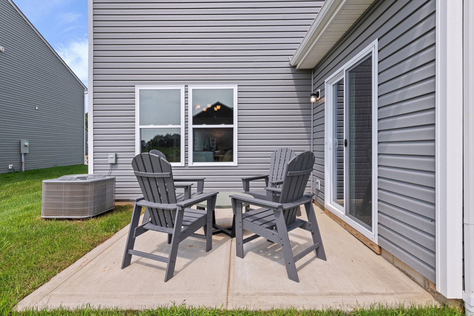 Outdoor patio with four gray Adirondack chairs on a concrete slab beside a modern house with gray siding.