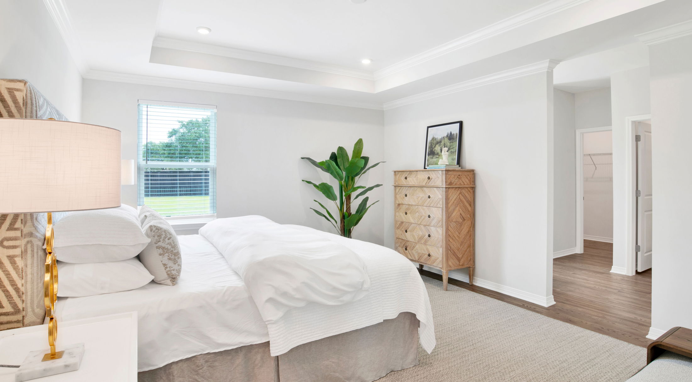 Bright and modern white bedroom with a cozy bed, wooden dresser, indoor plant, and natural light from a large window.