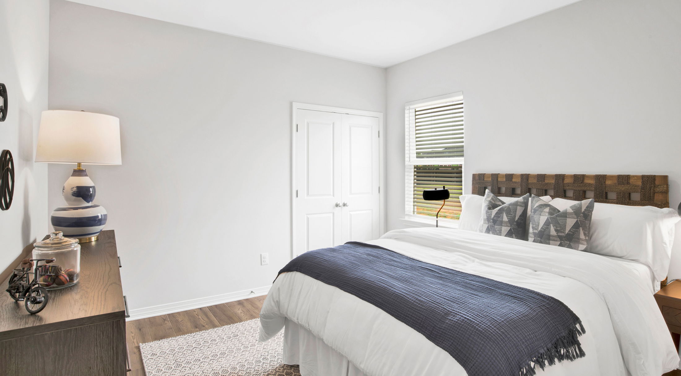A modern, bright bedroom with white walls, a wooden headboard, a neatly made bed featuring geometric accent pillows, and a deep blue throw blanket.