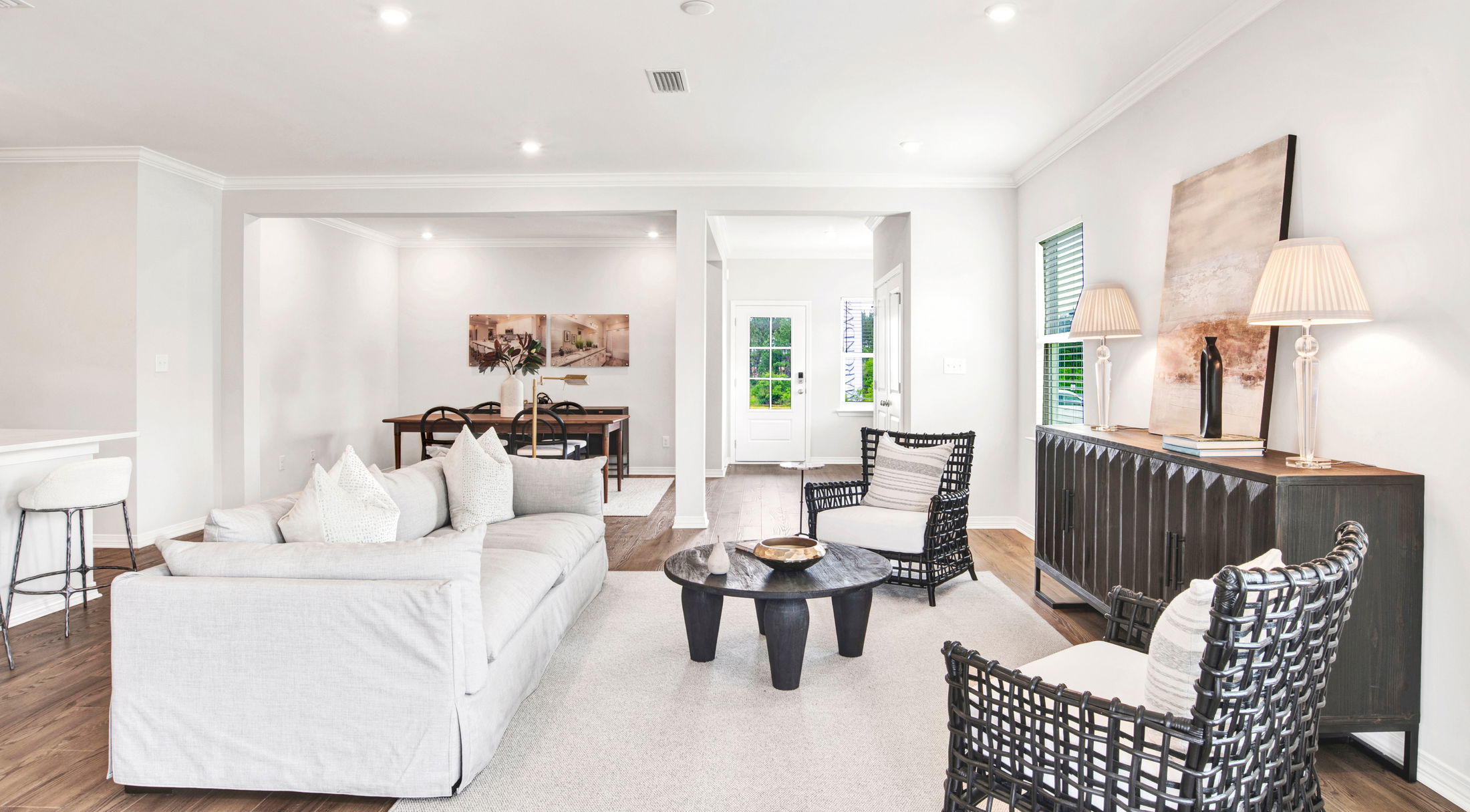 Modern living room with white walls, comfortable gray sofa, wicker chairs, and a sleek wooden dining area in the background.