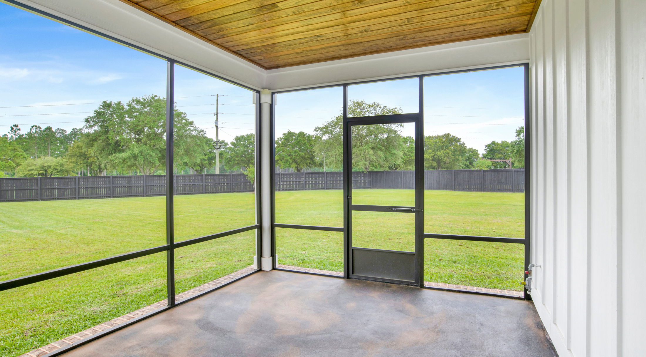 A screened-in porch with a wooden ceiling overlooking a spacious, well-manicured lawn enclosed by a black fence.