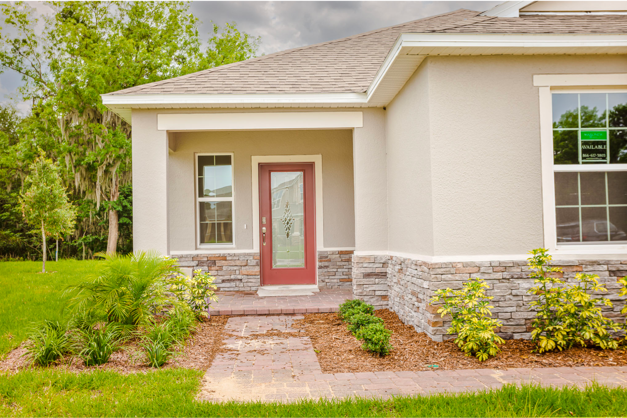 Front entrance of a modern house with a red door, stone facade, landscaped greenery, and a brick walkway.