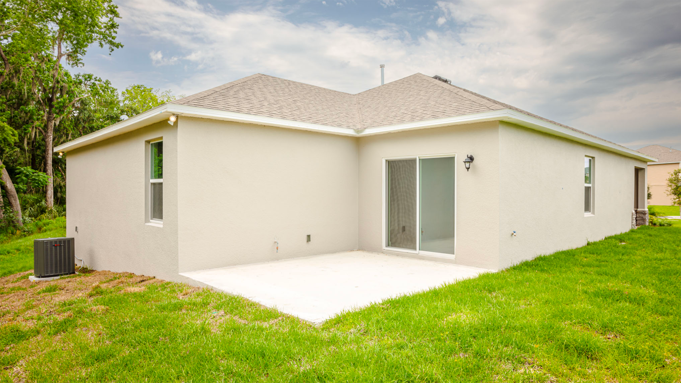 Backyard view of a modern beige house with a sliding glass door and green lawn, featuring a concrete patio and air conditioning unit.