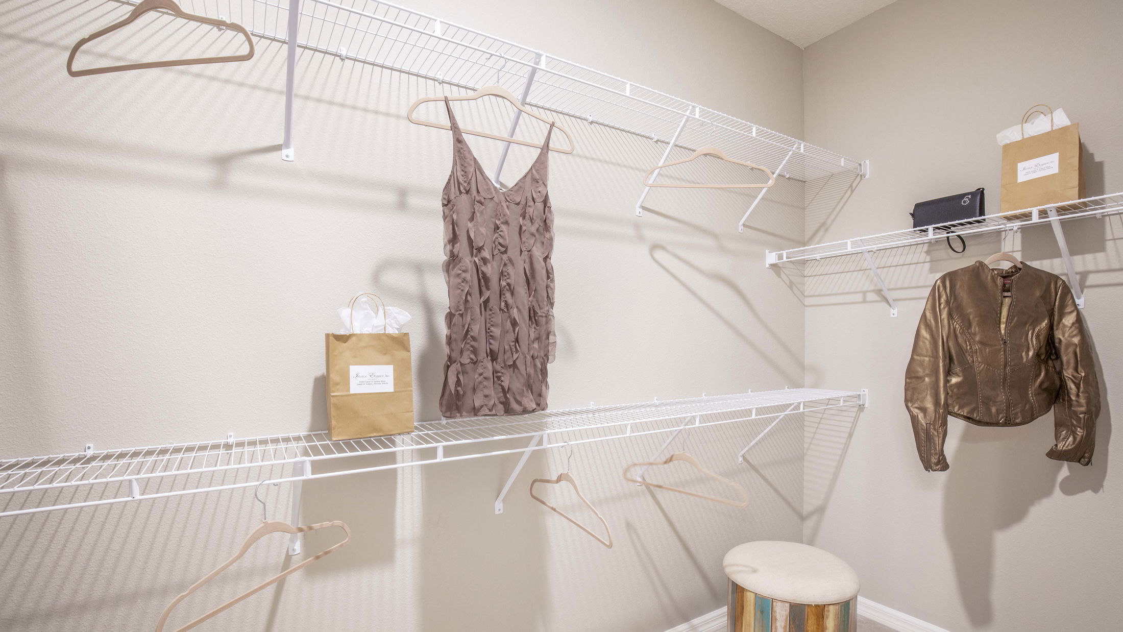 Empty white wire closet shelves with a brown sleeveless dress and leather jacket hanging, surrounded by shopping bags and a stool.